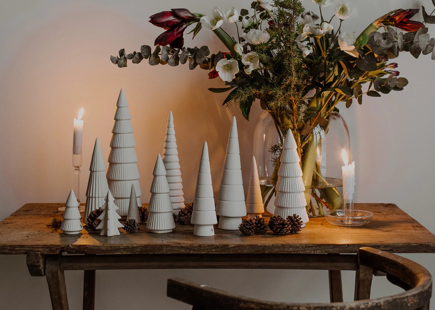 A wooden table decorated with various heights and shapes of white ceramic trees with a candle at each end and a bunch of winter foliage in a glass vase behind them