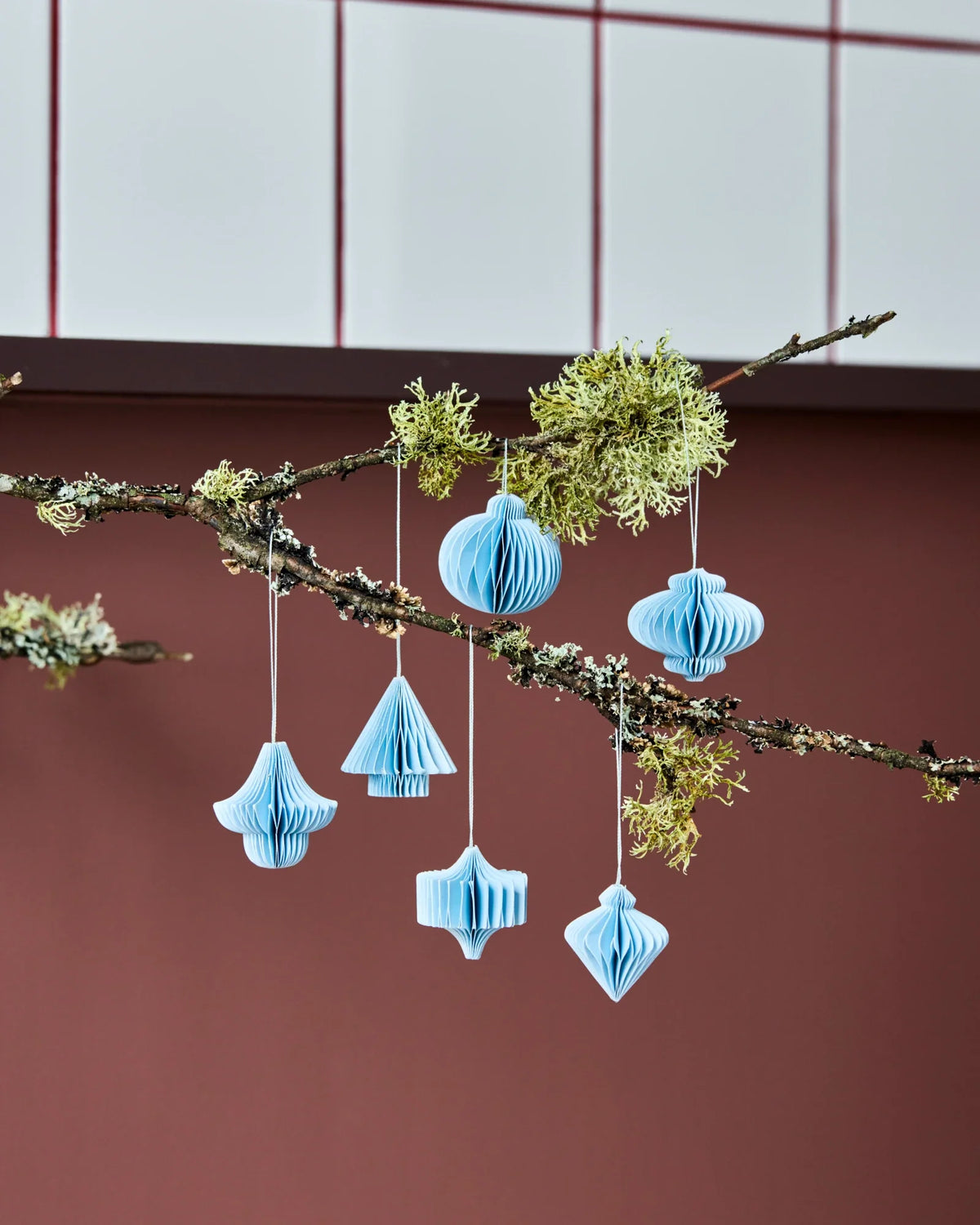 Light blue paper HSComb hanging ornaments by House Doctor in various shapes, suspended from a natural tree branch with green moss against a reddish-brown wall and white panelled ceiling.