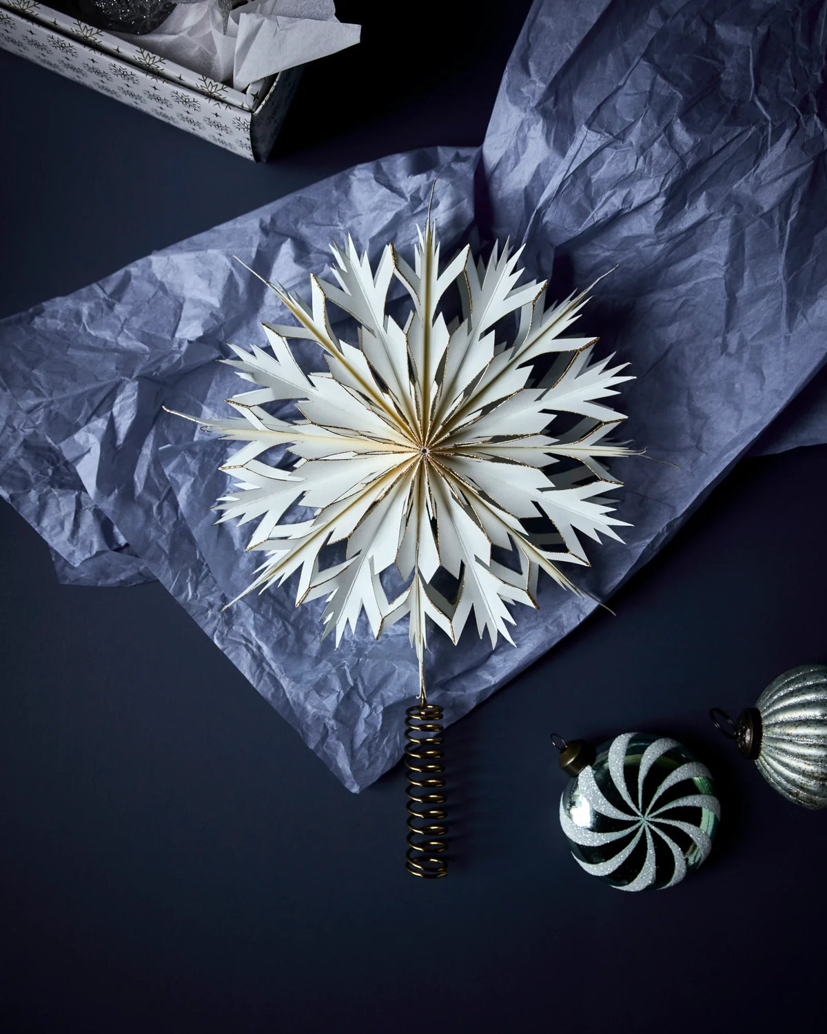 The HDAlsta white paper star Christmas ornament with gold accents and a dark spiral stem from House Doctor, styled on crumpled blue-grey tissue paper with holiday ornaments on a dark background.
