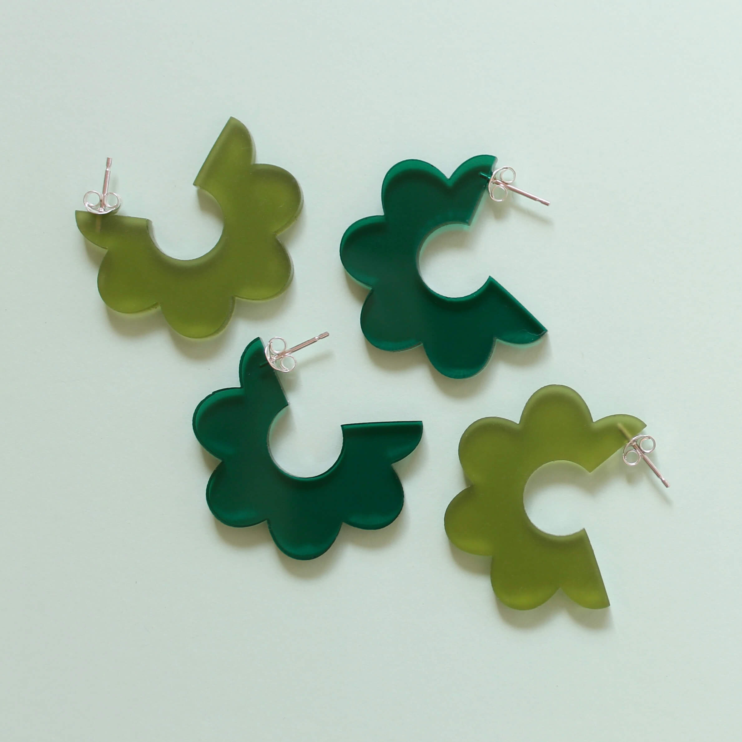 A pair of dark green and a pair of olive green scallop edged acrylic hoop earrings on a pale green background