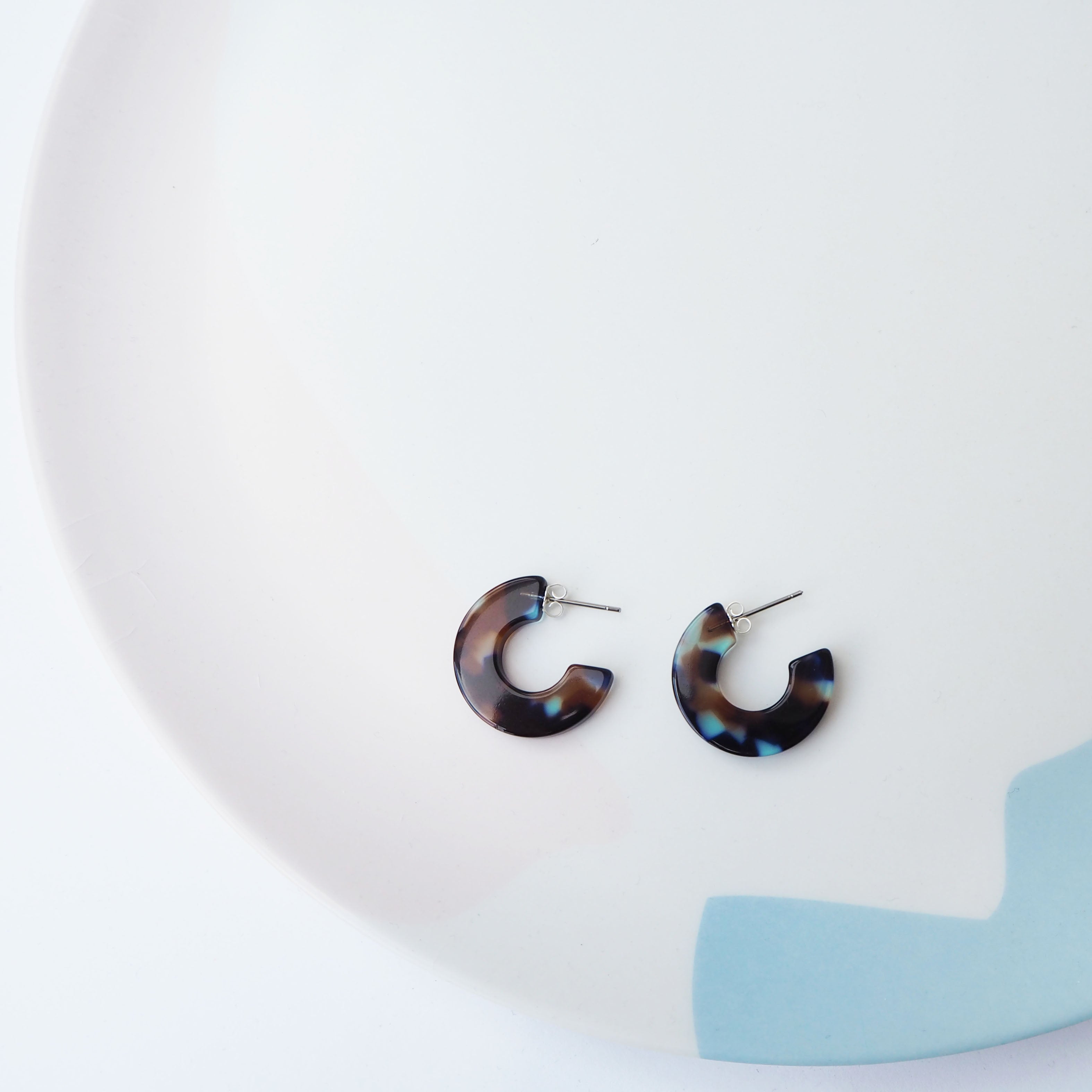 A pair of small acrylic hoop earrings in a mix of clear, blue and dark brown lying flat on a ceramic dish