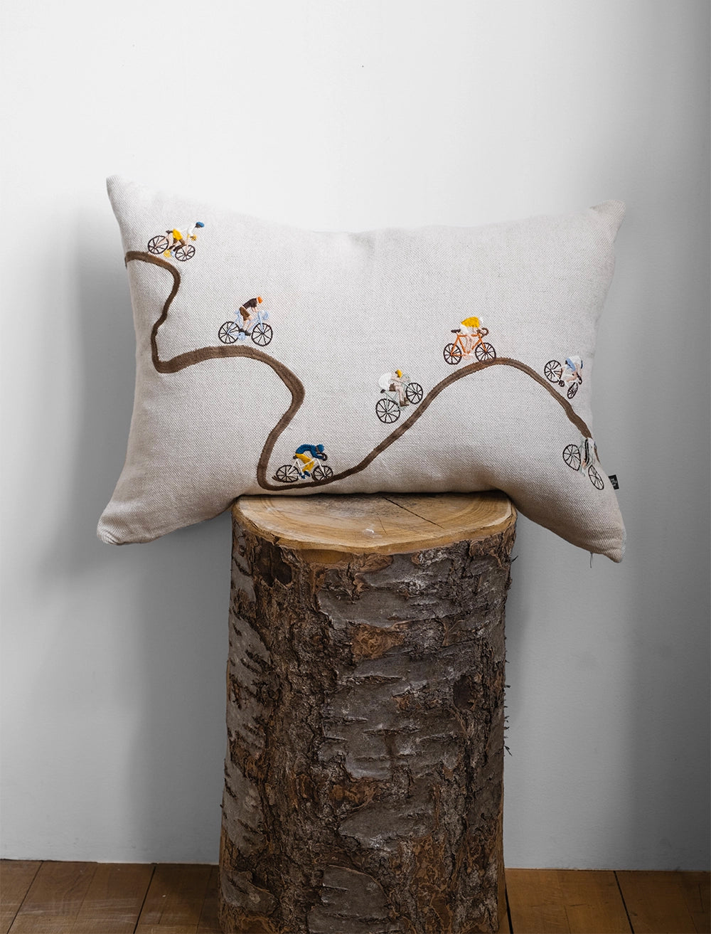 A rectangular cushion in natural linen with an embroidered path and cyclists from Fine Little Day balances on a cut log