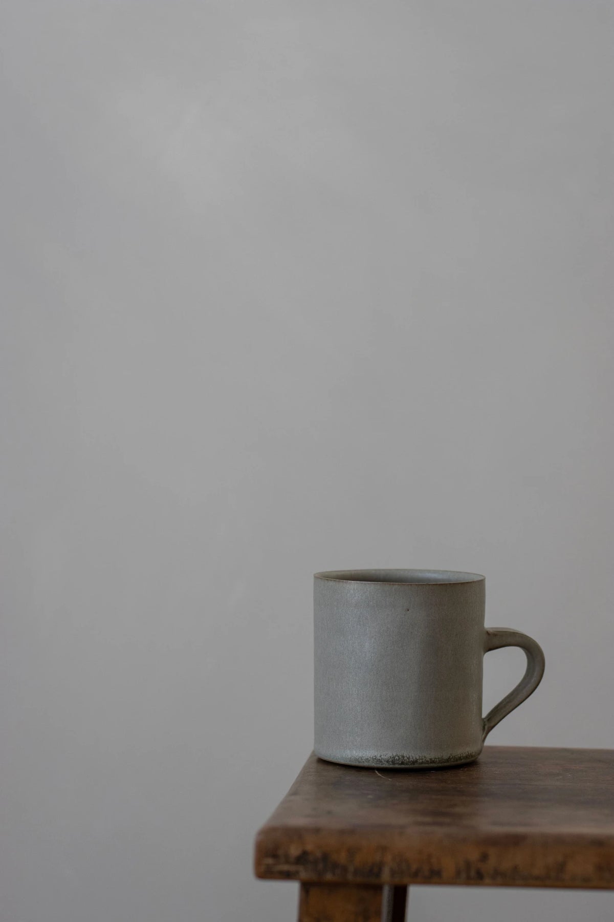 A Borja Moronta cylindrical cup in feather grey glaze sits on the corner of a rustic wooden table against a plain light grey wall.