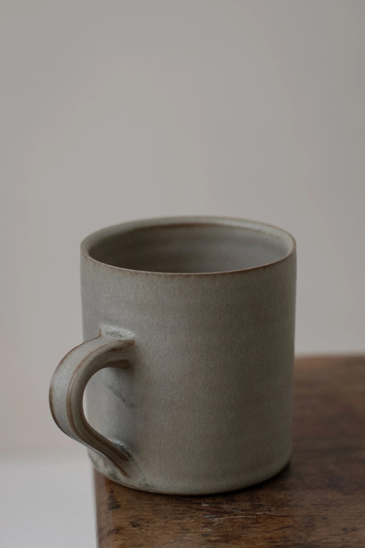 A Borja Moronta Cylindrical Cup in Feather Grey Glaze, resting on a rustic wooden surface against a neutral light grey background.