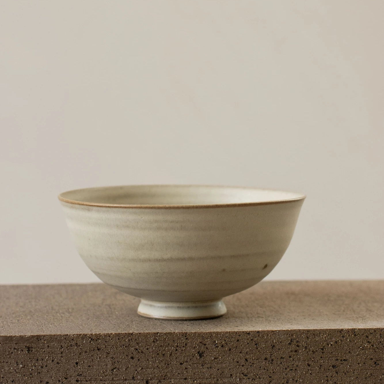 A Borja Moronta small footed bowl in warm white glaze, sitting on a dark stone surface against a warm beige background.