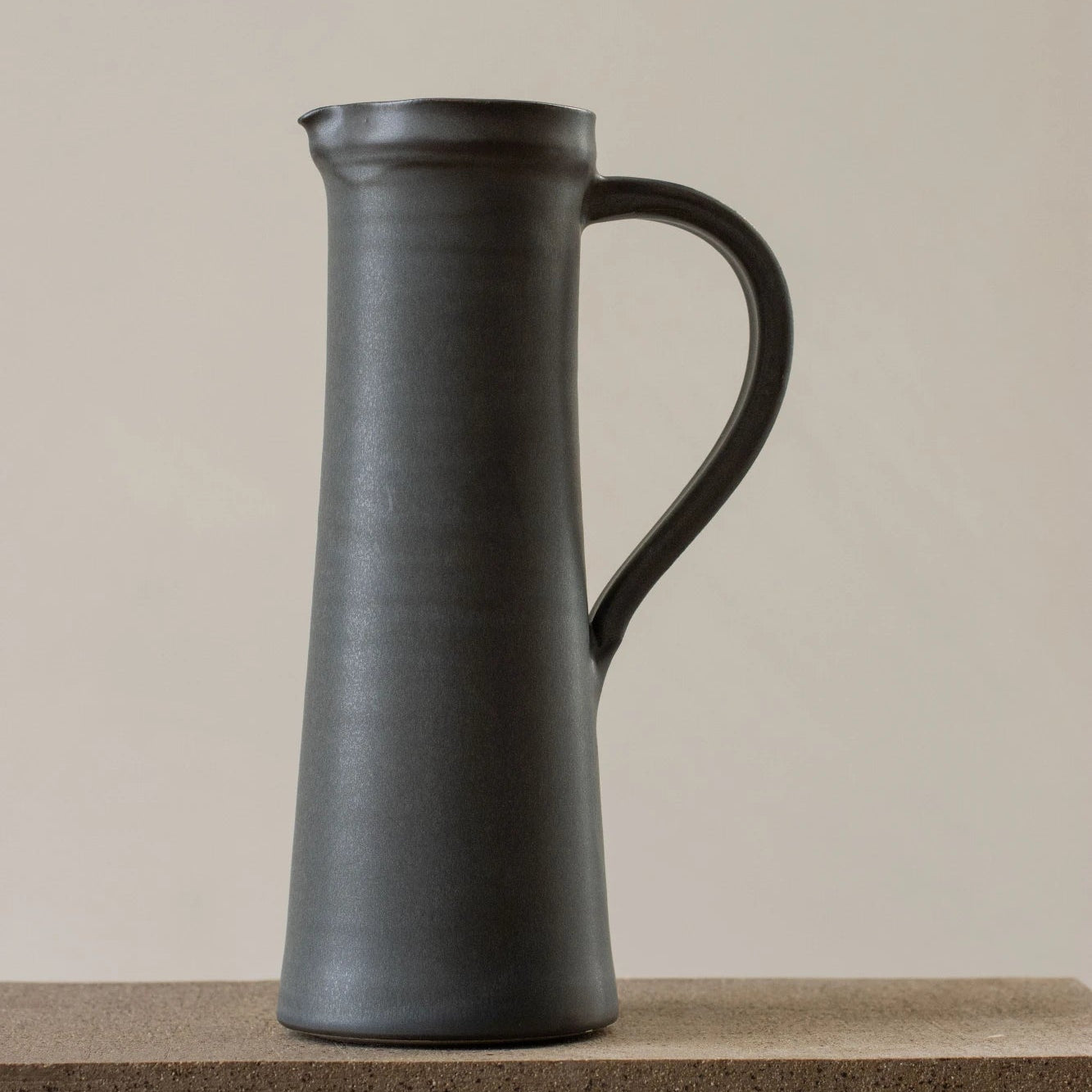 A matte dark gray ceramic pitcher with a curved handle, standing on a textured stone surface against a warm beige background.