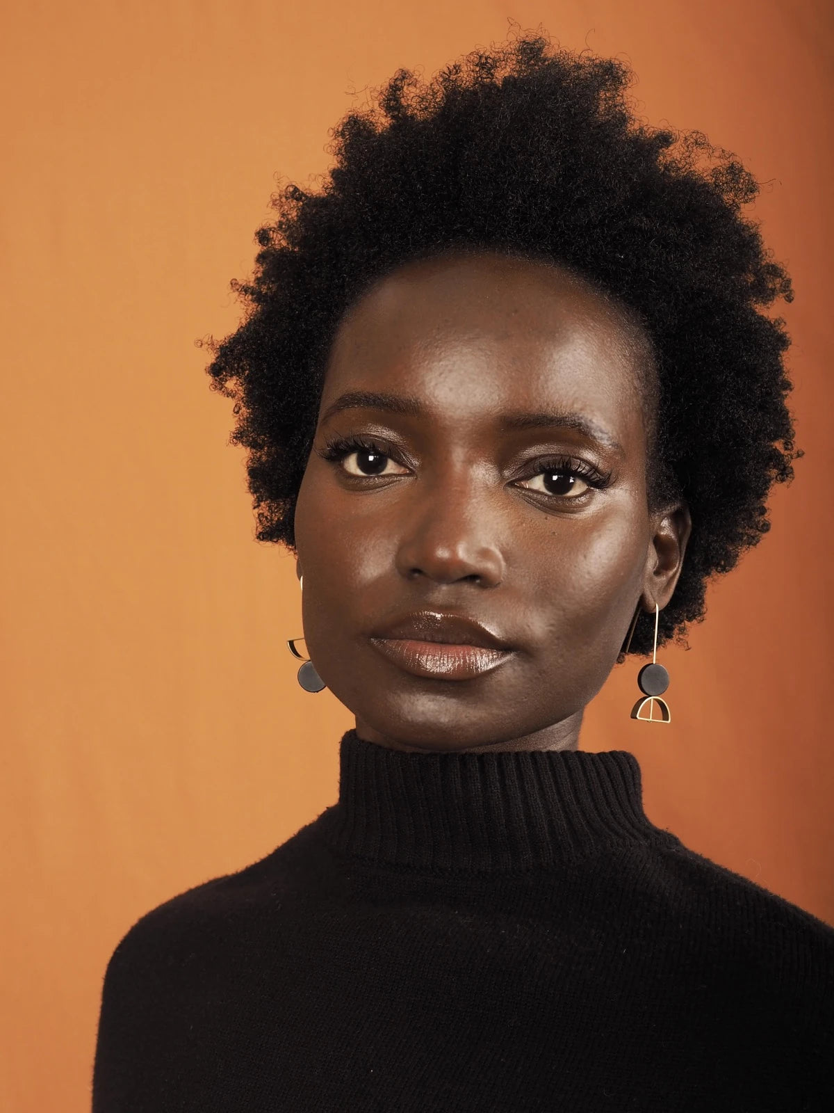 A close-up portrait of a woman with short, curly black hair wearing a black turtleneck sweater and Nucleo Brass and Black Upside Down Arch Earrings by brass+bold against a solid orange background.