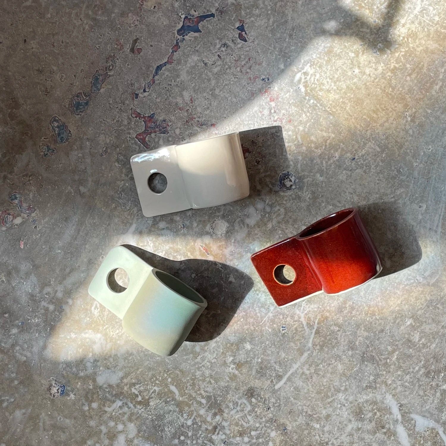 Brutes Ceramics Hopper Mugs in sand, green and burgundy lying flat on a grey stone floor with sunbeams on them