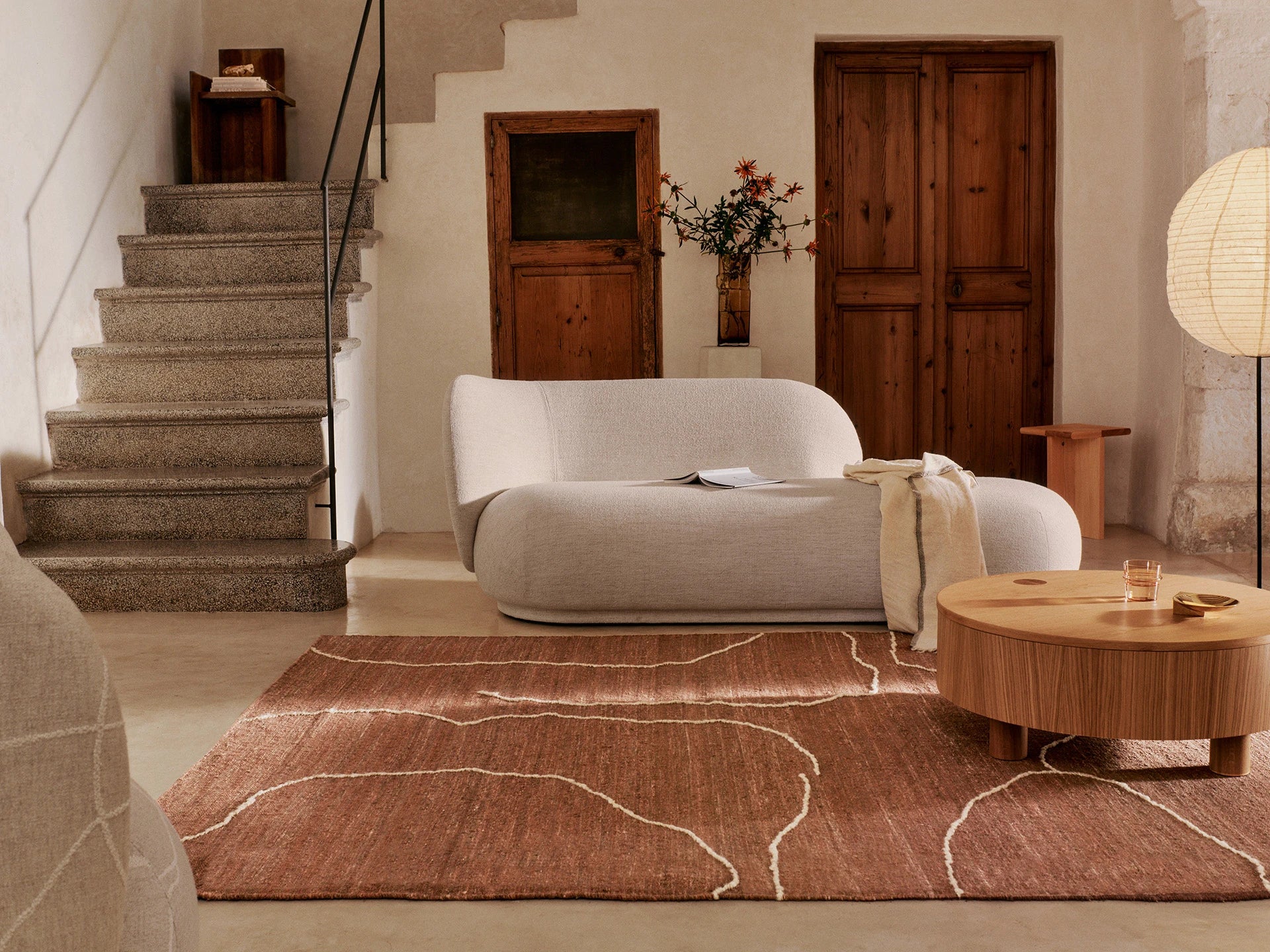 Ferm Living Rico Divan with left sitting backrest in off white boucle in a context image featuring a rust rug, wooden table and two wooden doors in the background