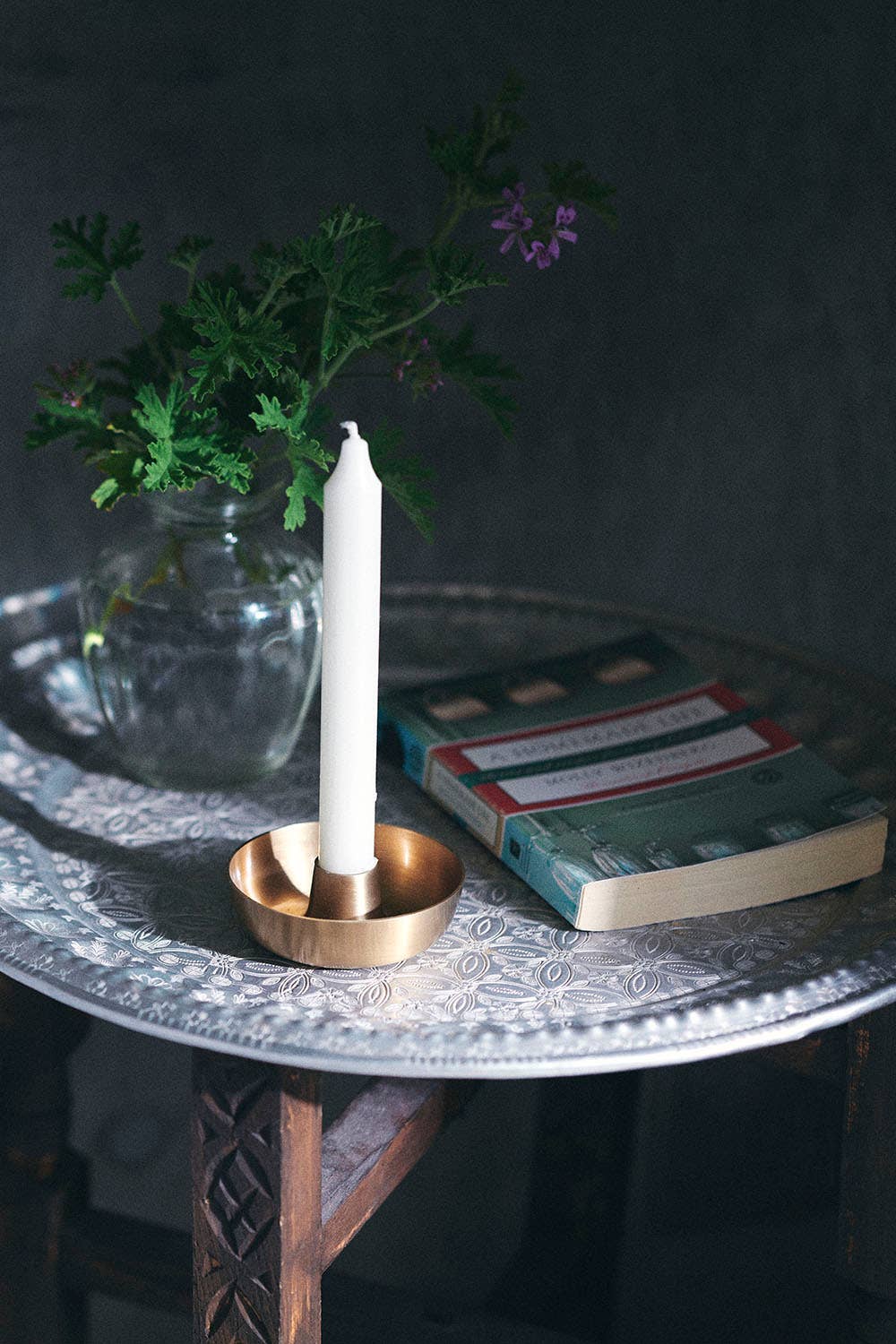 White candle in a brass holder on a decorative tray with a book and a geranium plant in the background