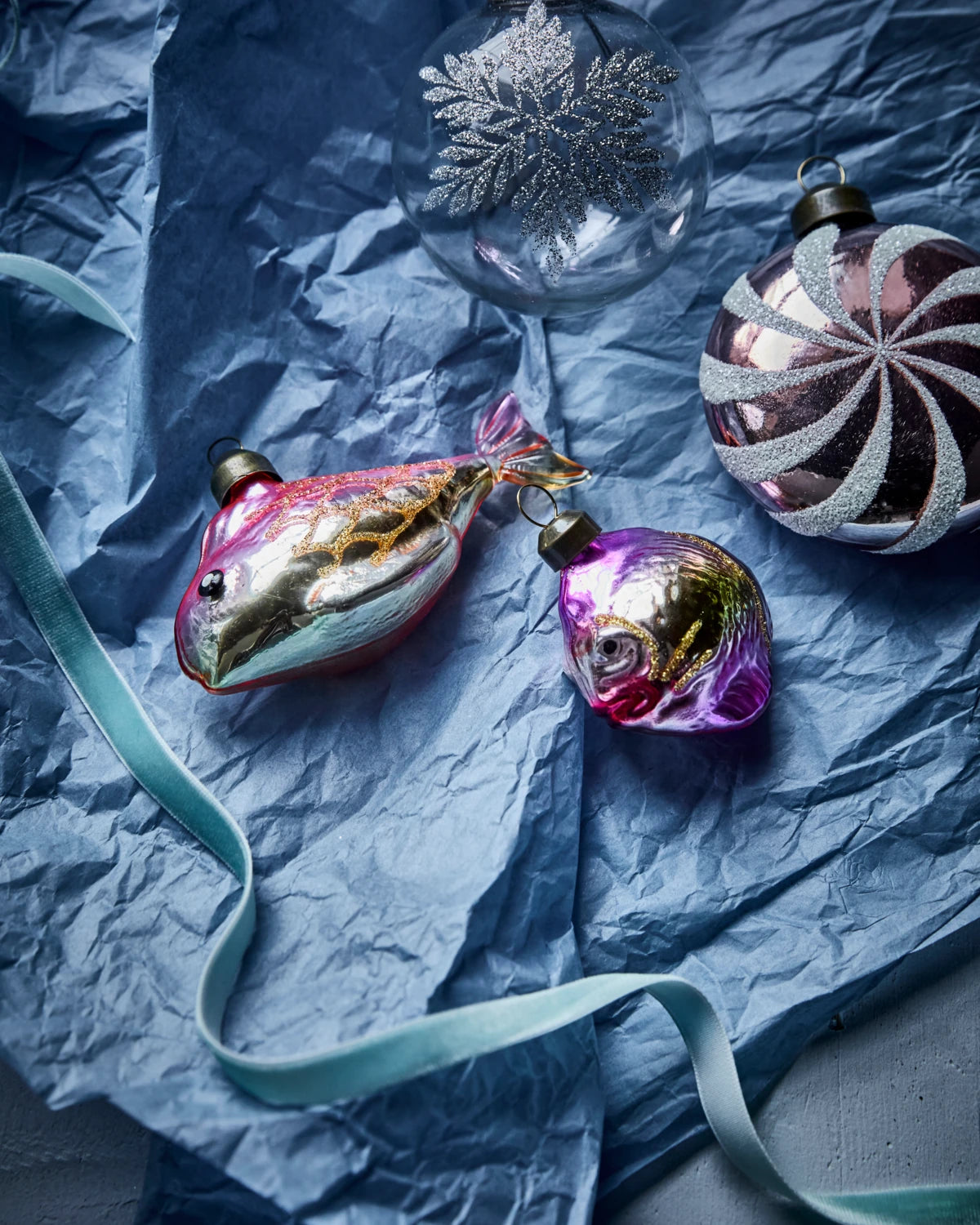 House Doctor Set of 4 Glass 'Pez' Hanging Ornaments in Gold & Pink lying with other ornaments on a textured blue paper surface