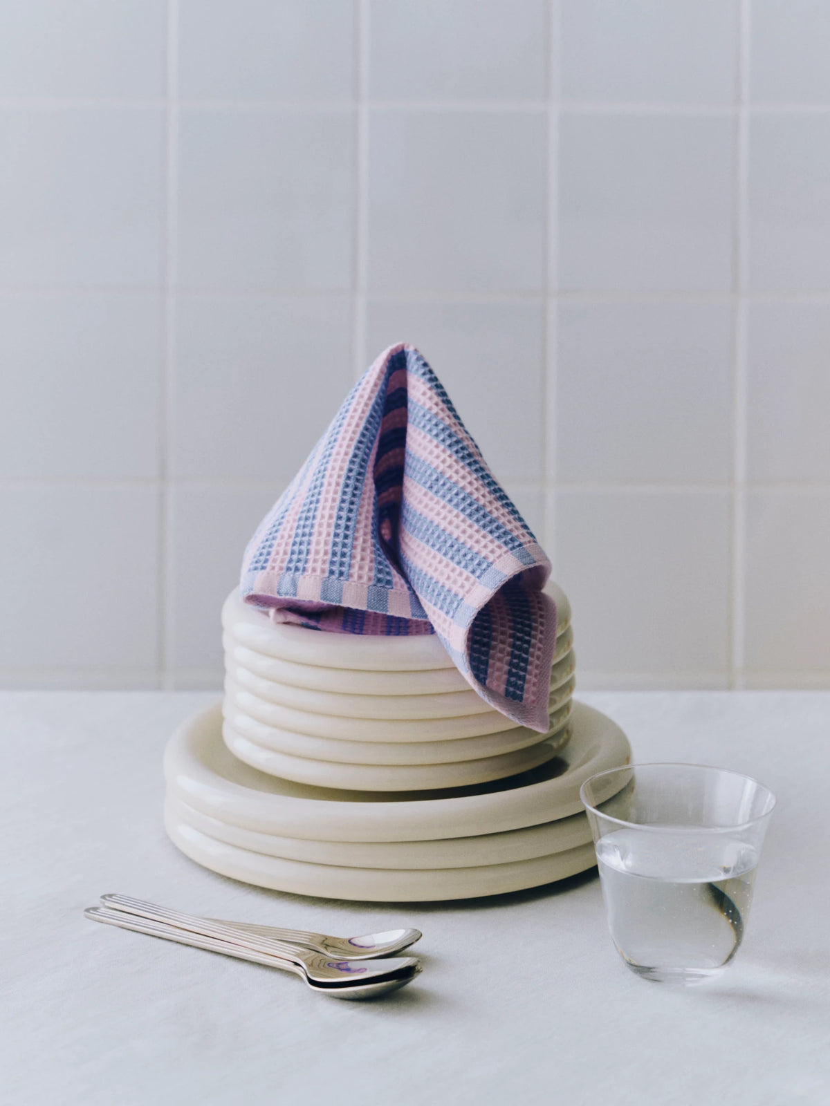 A stack of cream-colored ceramic plates with a purple and pink striped HAY Canteen wash cloth, set on a light marble countertop with silver spoons and a clear glass cup against a white tiled wall.