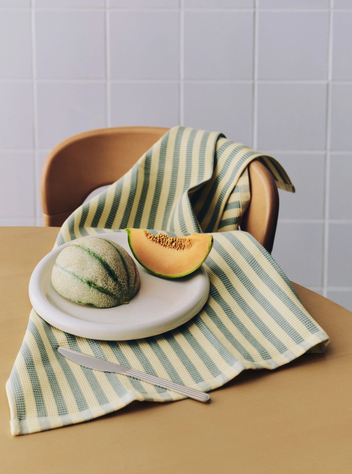 A green and yellow striped HAY Canteen kitchen towel draped over a wooden chair at a table, styled with a white plate holding a melon and a slice, a silver knife, and a light wood background.