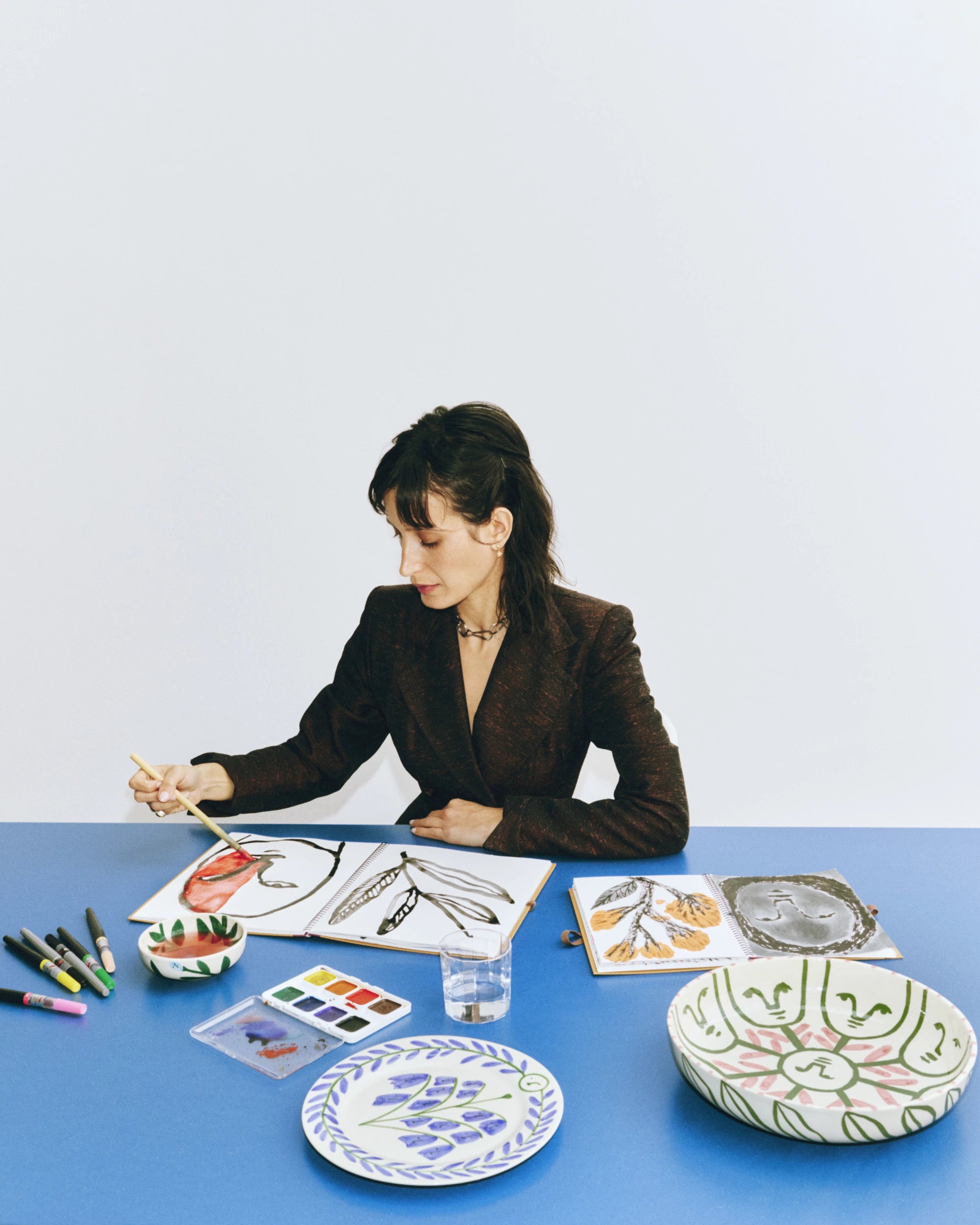 Artist Emma Kohlmann wearing a black jacket and sitting at a blue table, painting on a large notebook with 2 tableware pieces from the La Pittura collection next to her