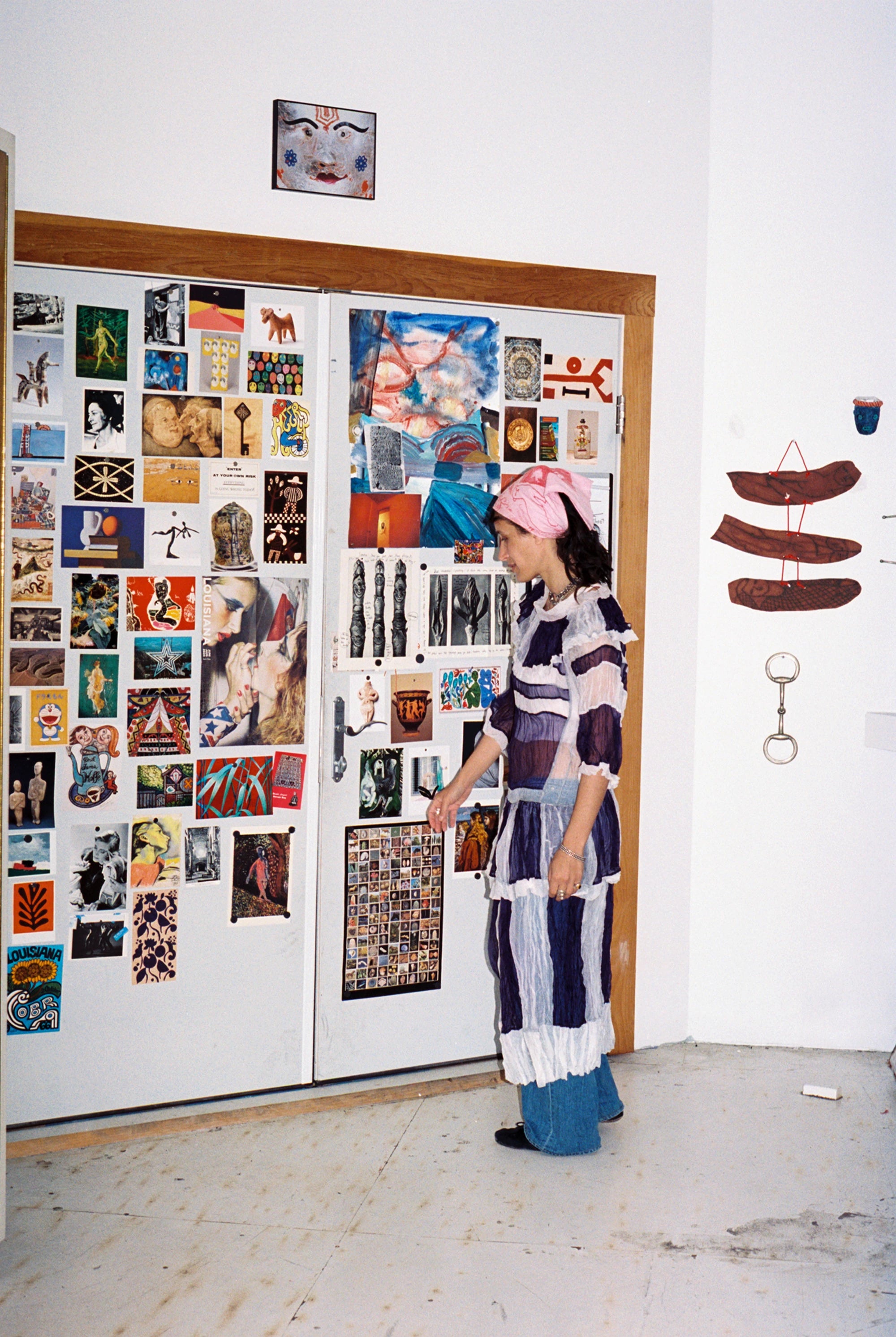 Artist Emma Kohlmann stands in front of a wall in her studio covered in inspirational images and art