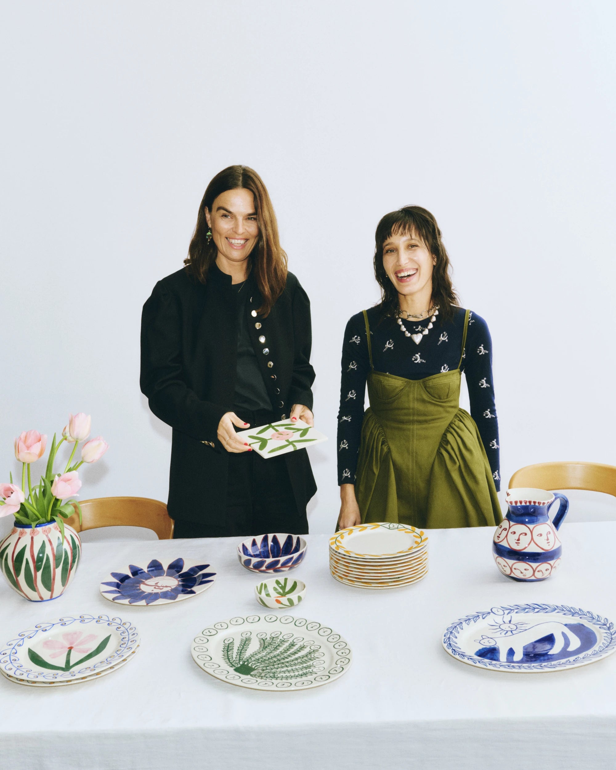 Mette HAY wears all black and stands next to artist Emma Kohlmann who wears a black top and olive green dress, They stand in front of a table with a white table cloth covered in piles of the La Pittura collection tableware