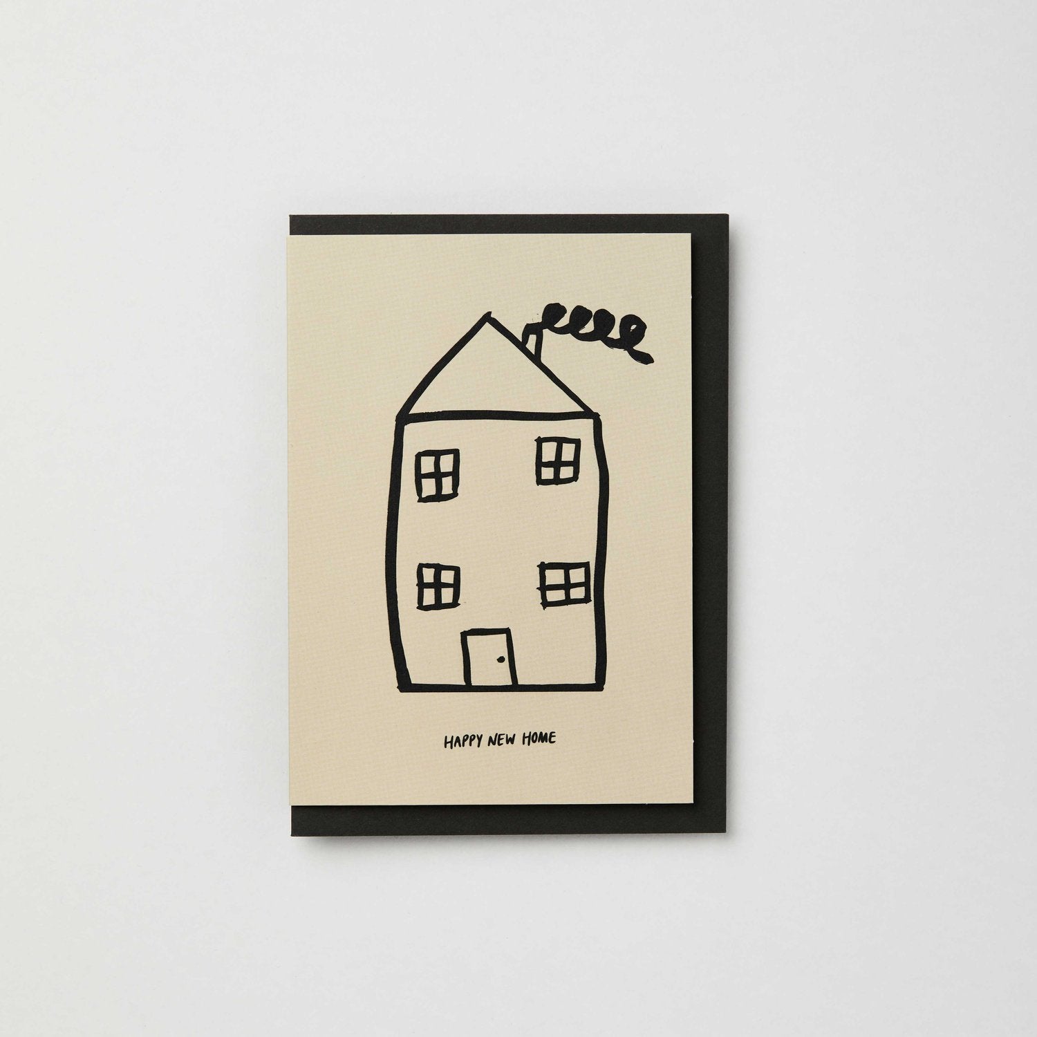 A plain white background with centred greetings card featuring a black illustrated house ans the text Happy New Home underneath, with a charcoal envelope, by Kinshipped