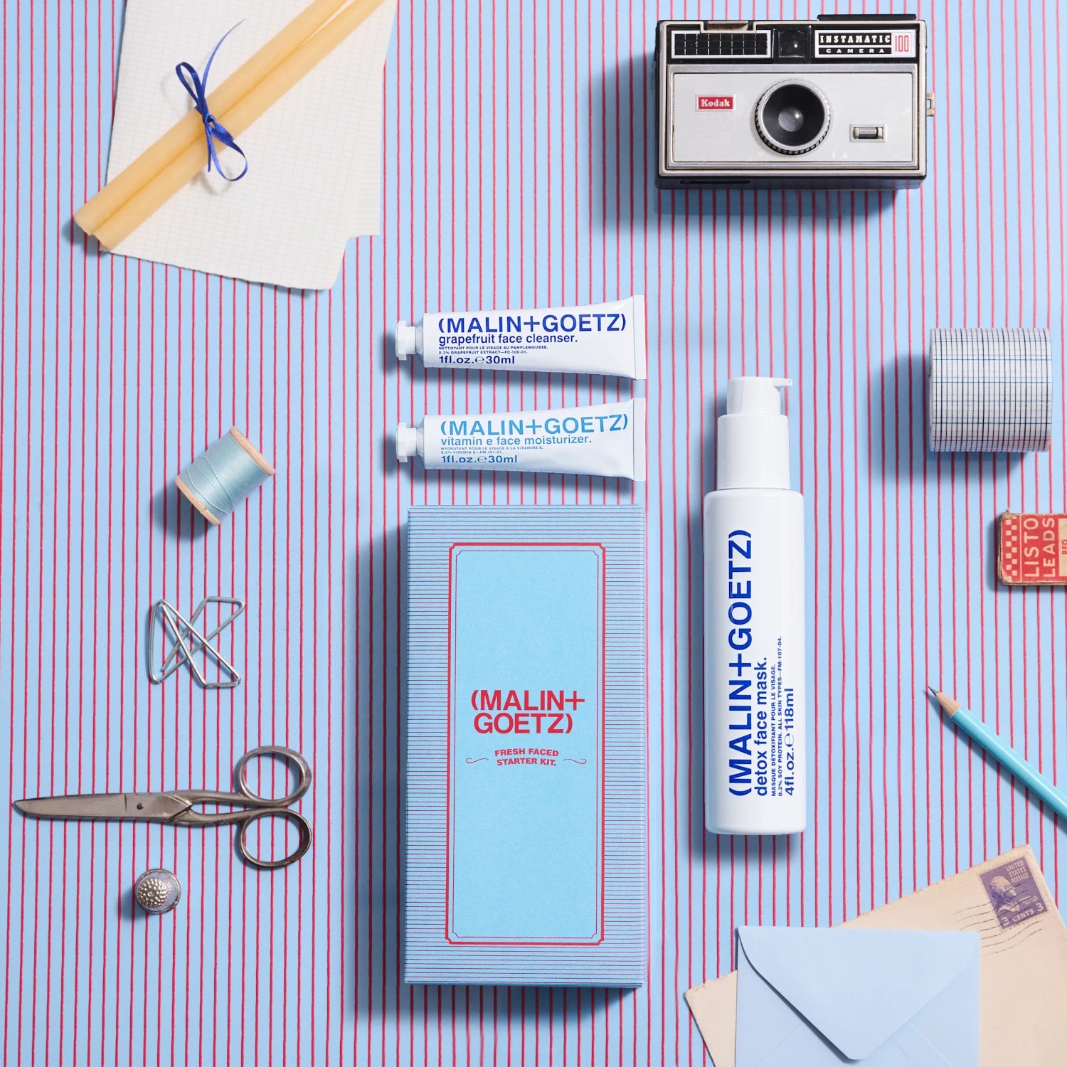 Malin and Goetz Fresh Faced Start Kit 2024 as a flatlay on a blue and red striped background with various stationery items surrounding them