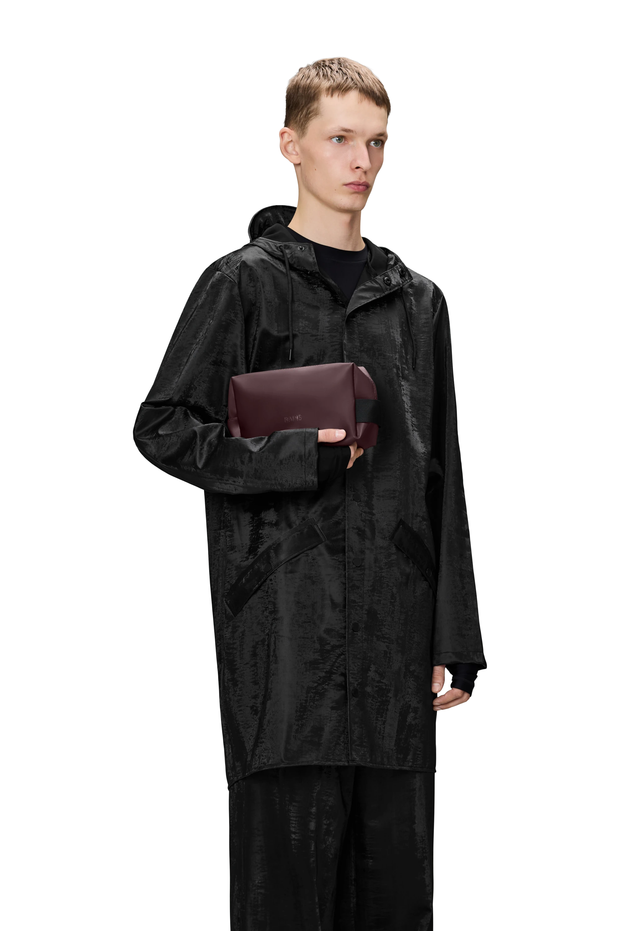 A model wearing a black waterproof jacket and trousers holdings the Rains Small Wash bag in Depth brown shade