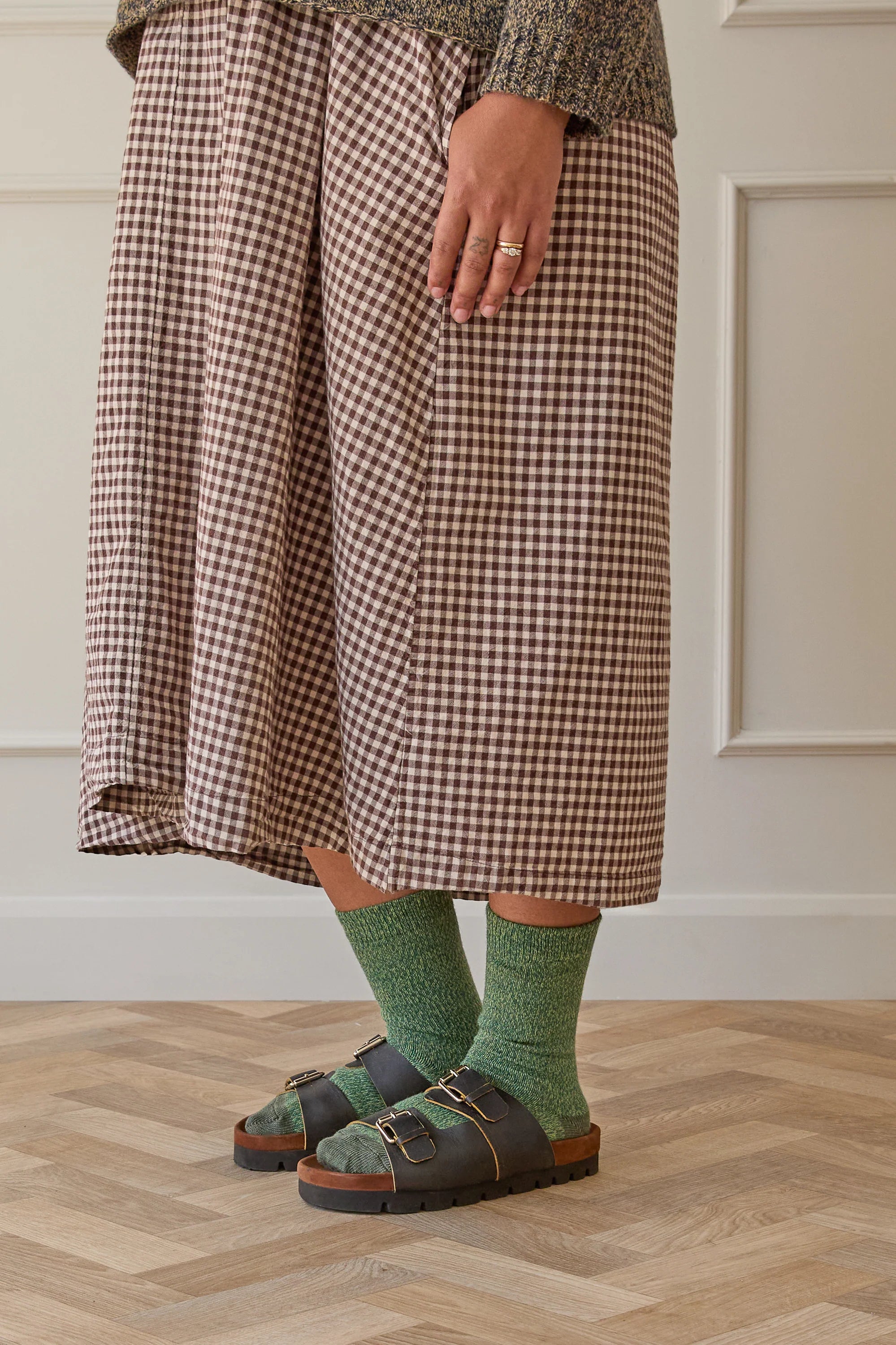 Person wearing a plaid skirt, green socks, and sandals on a wooden floor.
