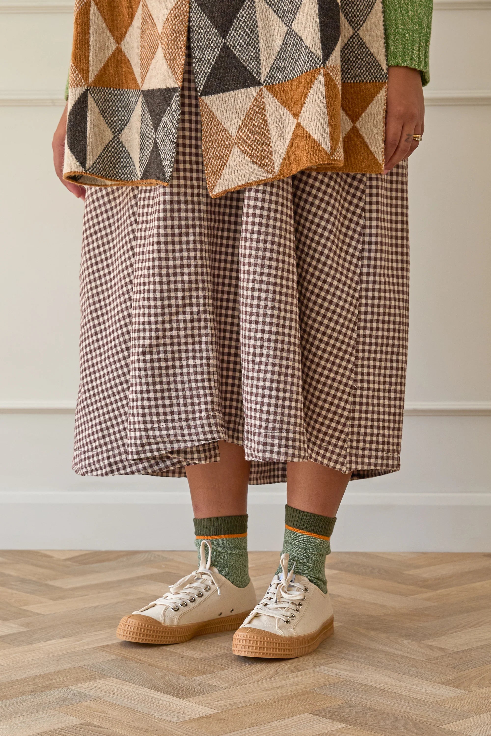 Person wearing a patterned coat, plaid skirt, and green socks with white shoes on a wooden floor.