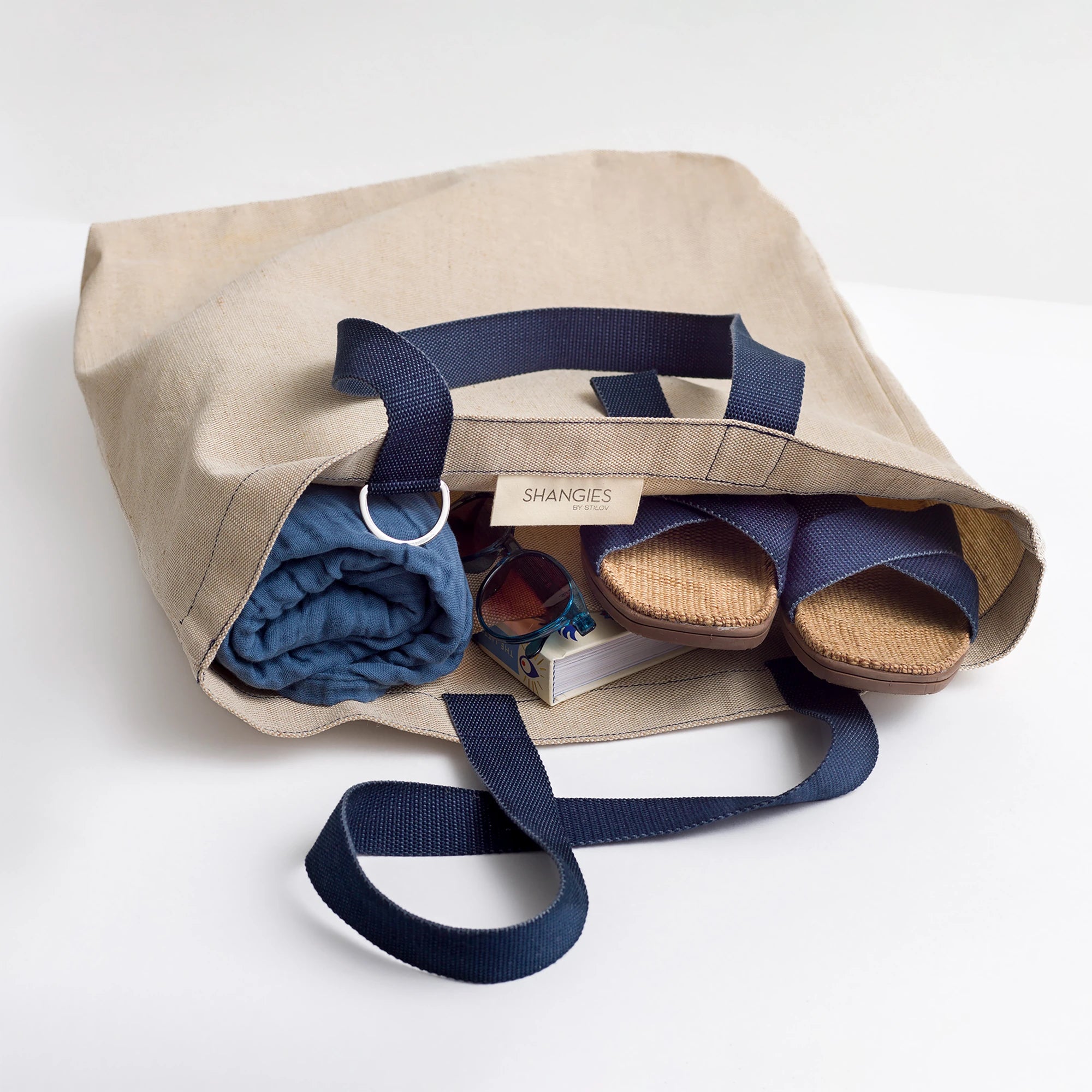 Beige tote bag by Shangies with Midnight Blue handles and strap, containing folded blue fabric, sunglasses, and books on a white background | Lifestory