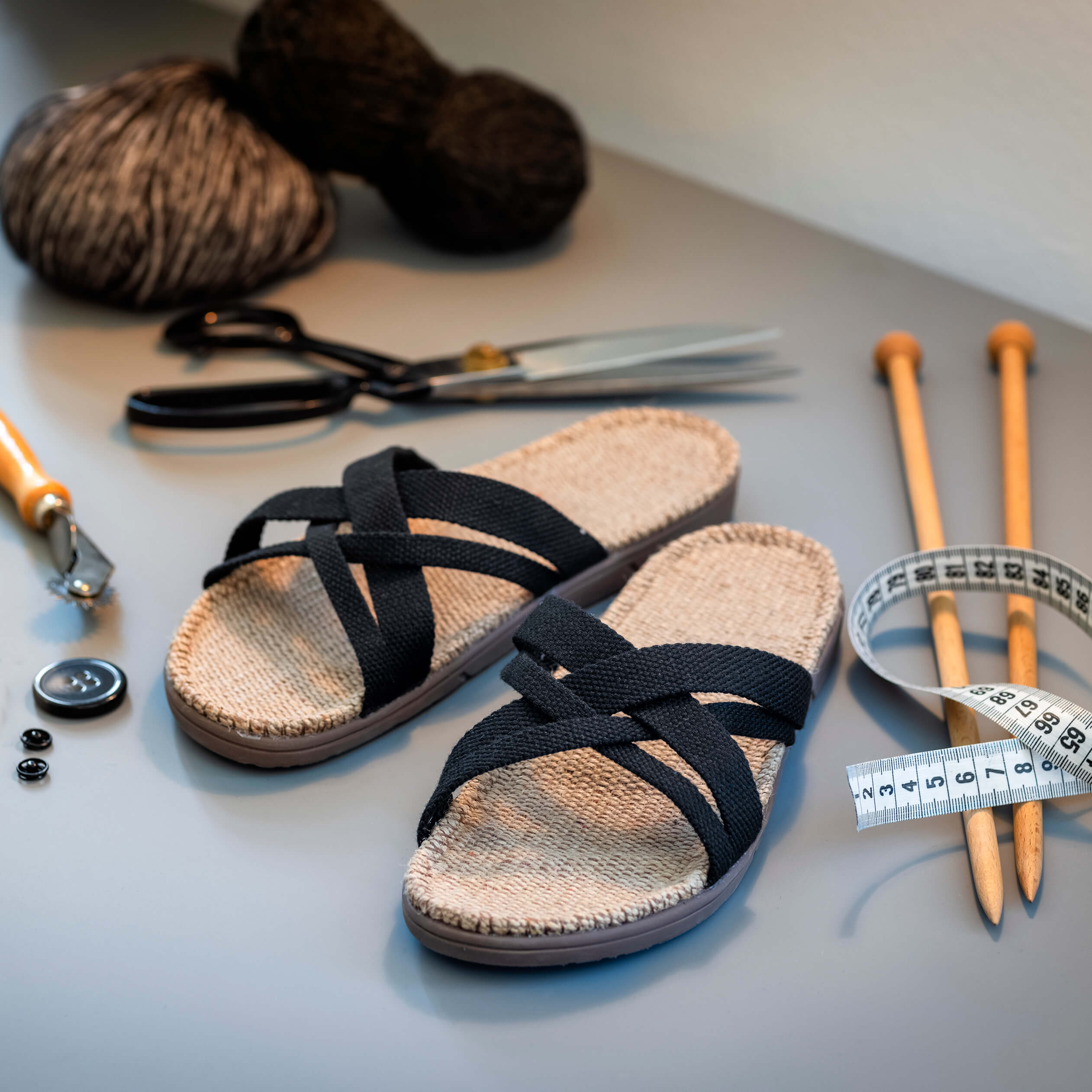 A pair of black strapped jute sandals sit next to knitting supplies on a grey background