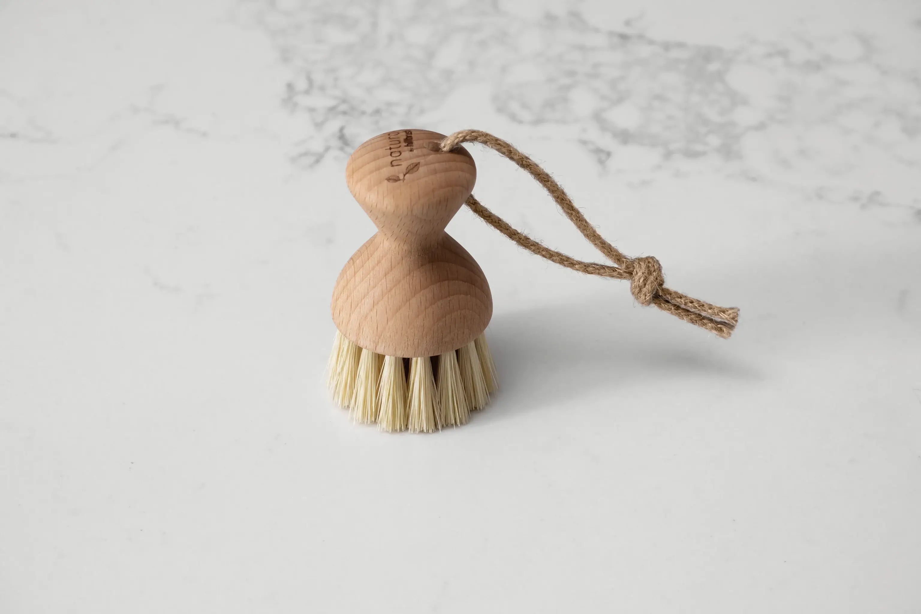 A small round wooden vegetable brush with compact handle and hanging loop sits on a marbled surface. Made by UK makers Hillbrush