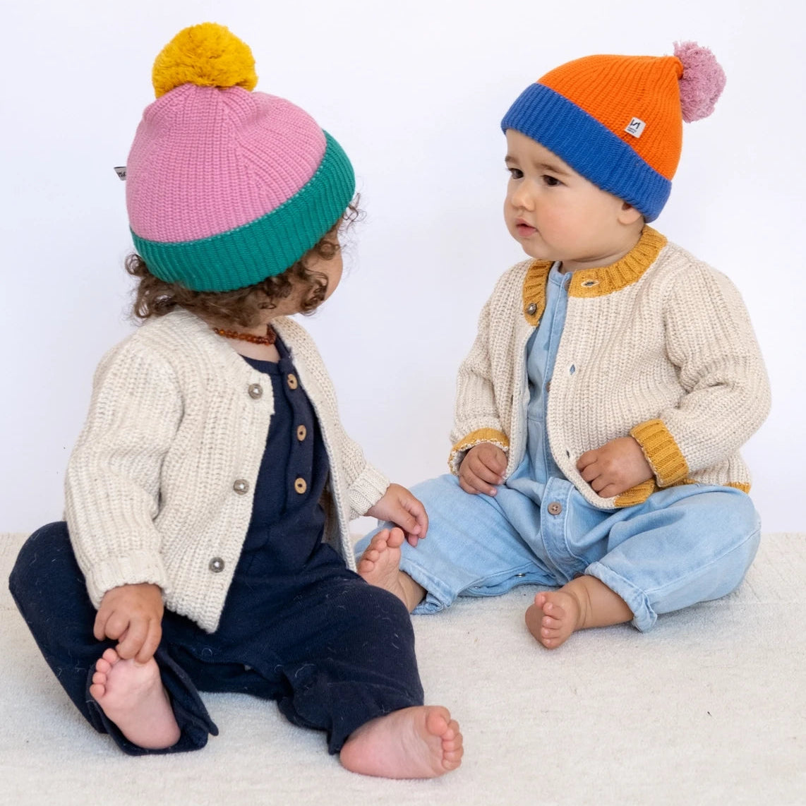Two children wearing colorful knit hats sitting on a white surface.