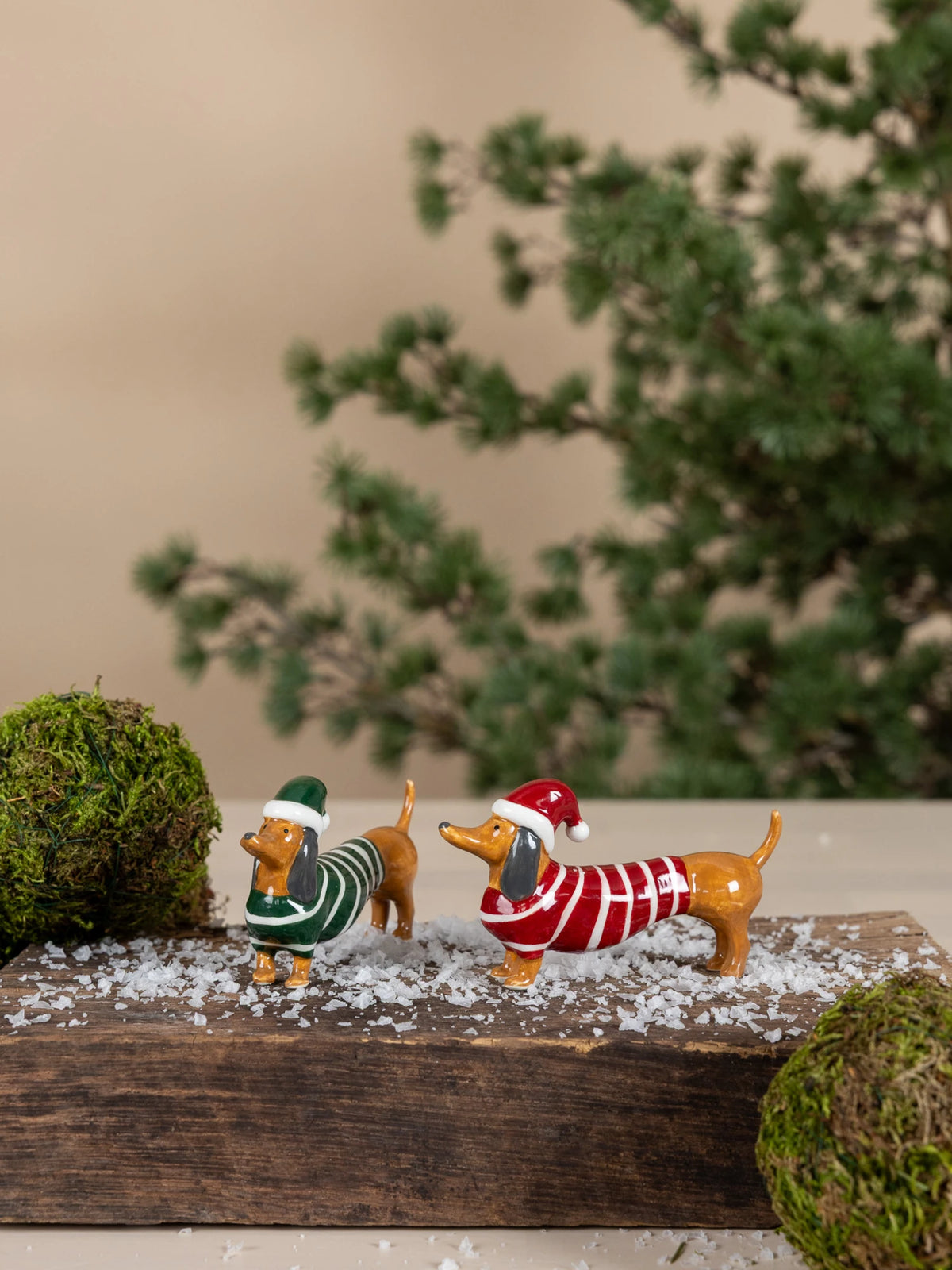 Two small dachshund Albin figurines by Storefactory dressed in holiday sweaters and Santa hats, standing on a wooden base with faux snow, moss balls, and a blurred evergreen background.