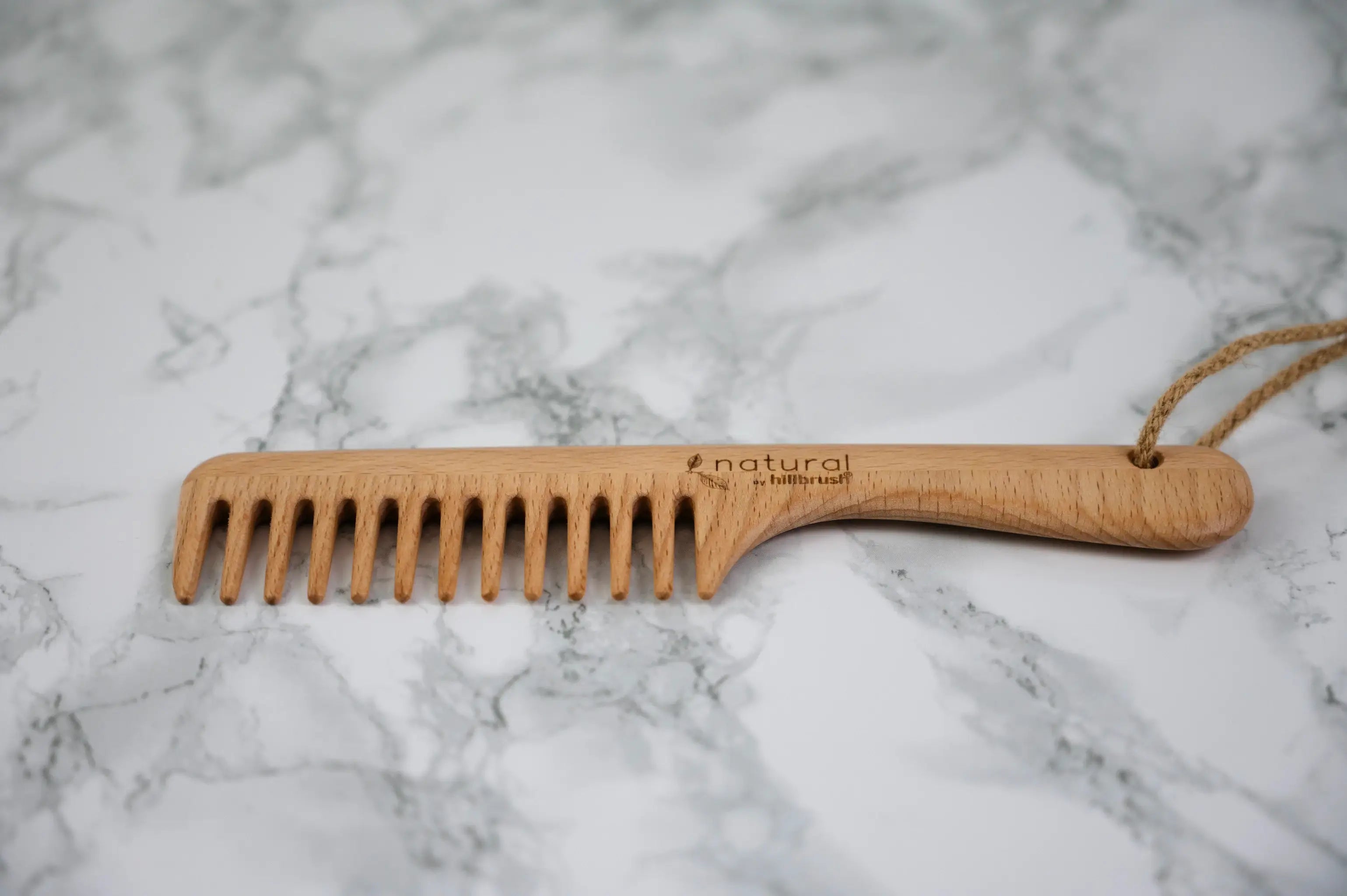 A wide toothed wooden comb with hanging loop lies flat on a marble surface. Product made by UK makers Hillbrush