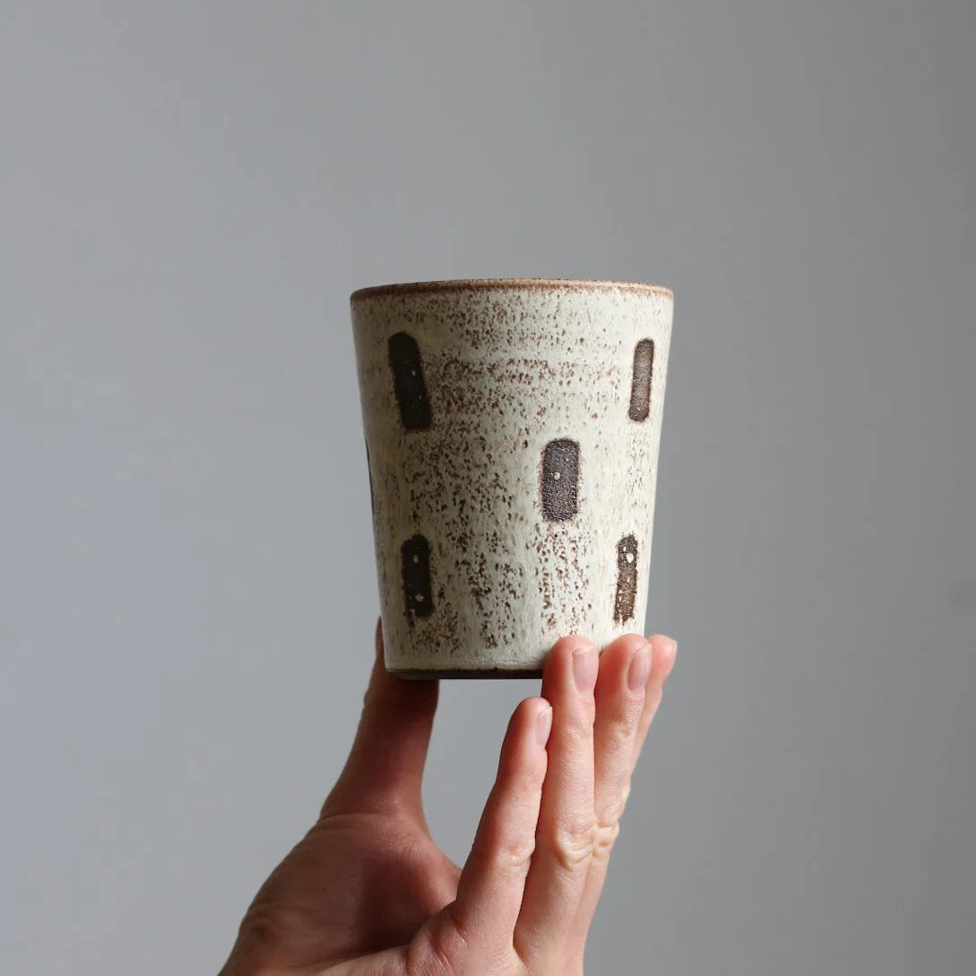 A hand holding a small cylindrical ceramic cup with a speckled beige and white glaze and dark brown rectangular accents, set against a plain gray background.