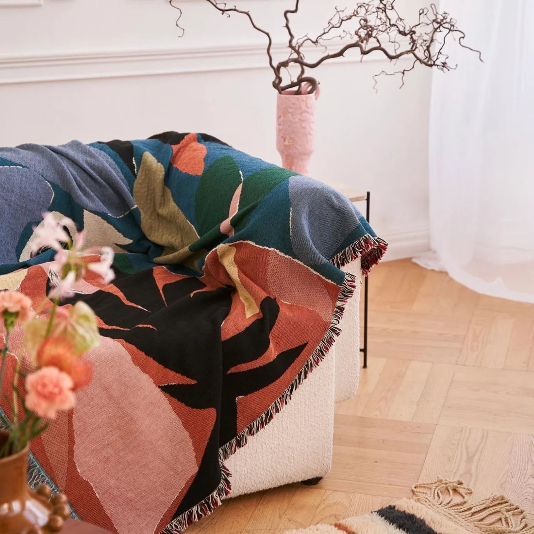 Colourful illustrated throw blanket in orange, greens and black draped over a sofa arm with a vase containing branches behind it