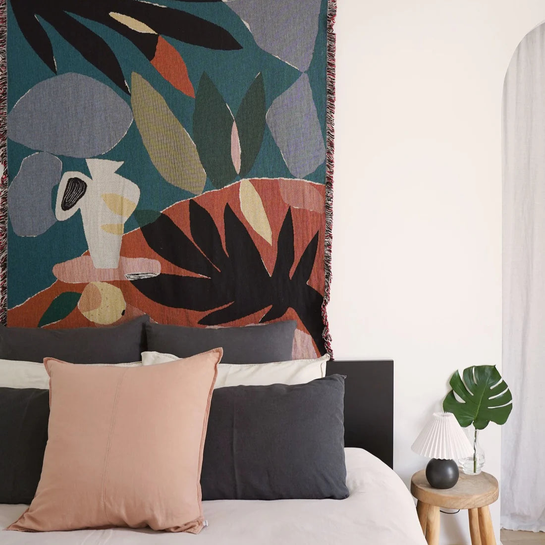 Bedroom scene featuring illustrated throw blanket in orange, greens and black hanging on a wall behind a bed