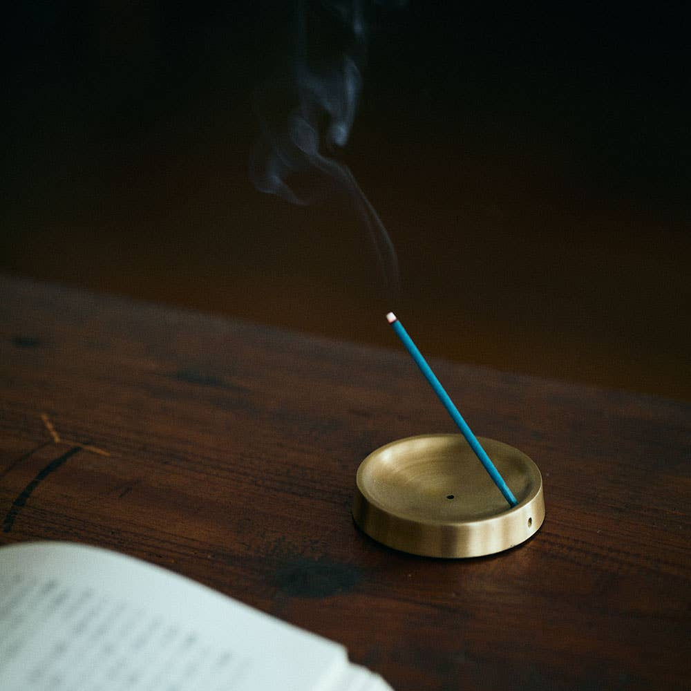 Incense stick burning in a brass holder on a wooden surface with an open book nearby.