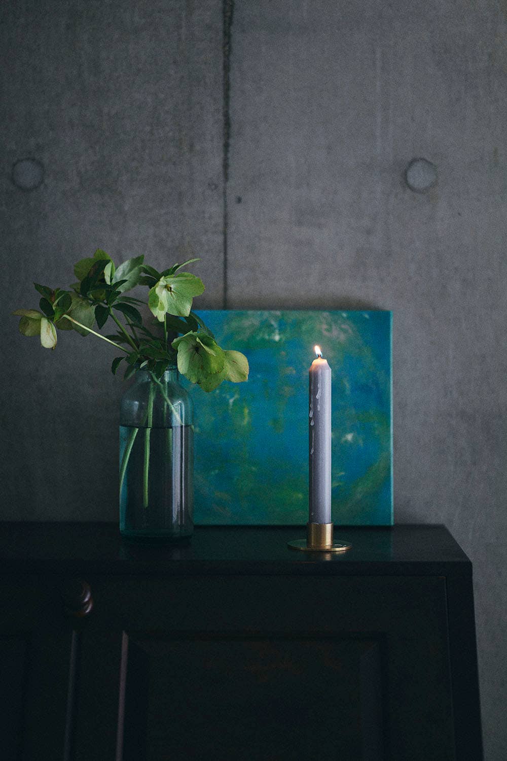 Vase with green leaves, blue abstract painting, and lit candle on a dark surface against a gray wall.