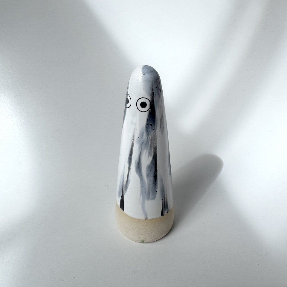 Dani Studio Arhoj ghost with a white and black marbled body, large eyes, and a natural beige base, standing on a white surface under dramatic side lighting.
