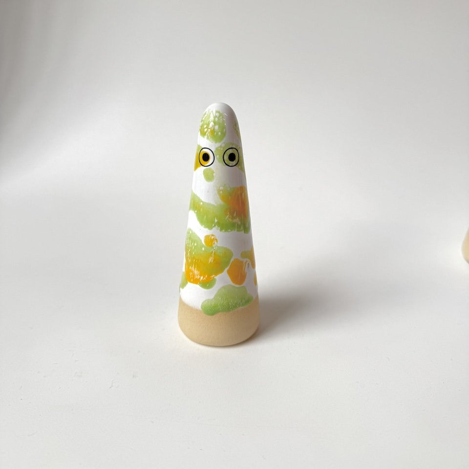Liam Studio Arhoj ghost on a white surface against a light grey background. The ghost has a natural beige clay base, a white upper body, and is glazed with hand-painted green and yellow patterns.