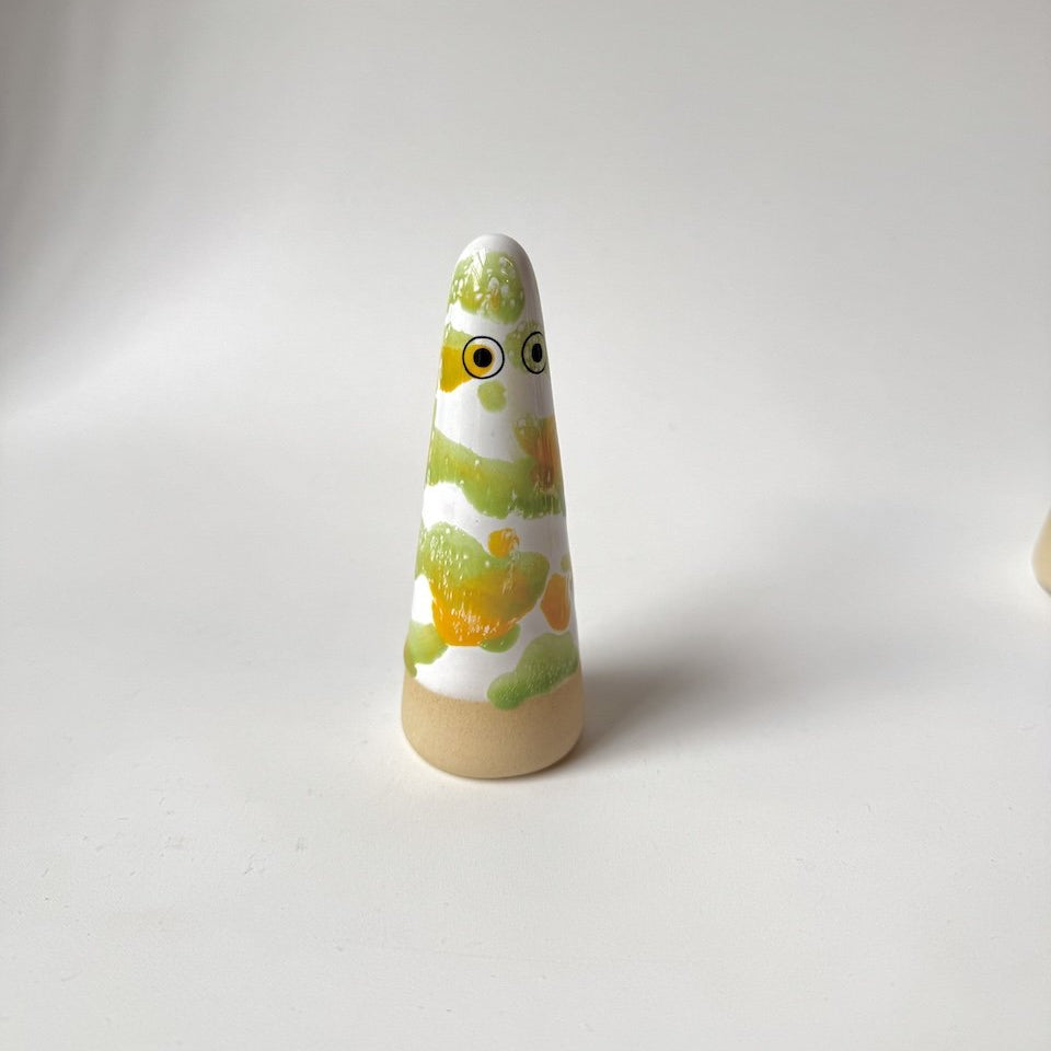 Liam Studio Arhoj ghost on a white surface against a light grey background. The ghost has a natural beige clay base, a white upper body, and is glazed with hand-painted green and yellow patterns.