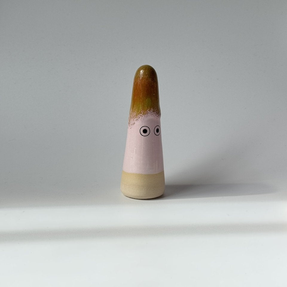 Elowen a Studio Arhoj ghost, featuring a light pink body with a brown and green glazed top, and painted black-and-white eyes, set against a plain light gray background.