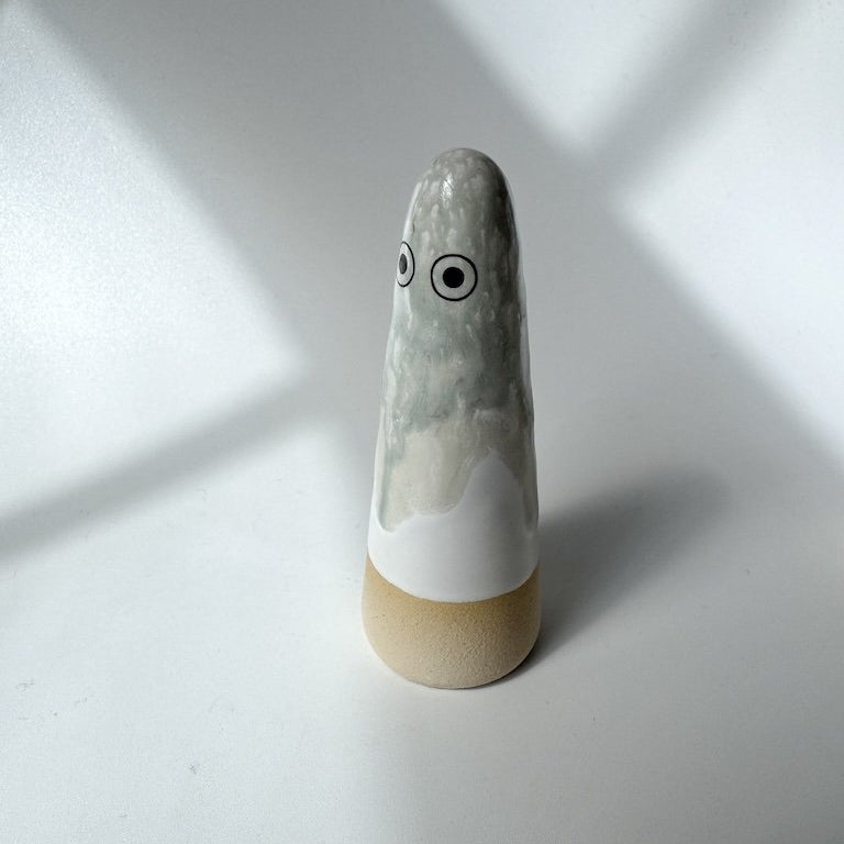 Jayne Studio Arhoj ghost with a glossy white and grey marbled upper body, black circular eyes, and a matte beige base, photographed on a light grey surface with soft window-light shadows.