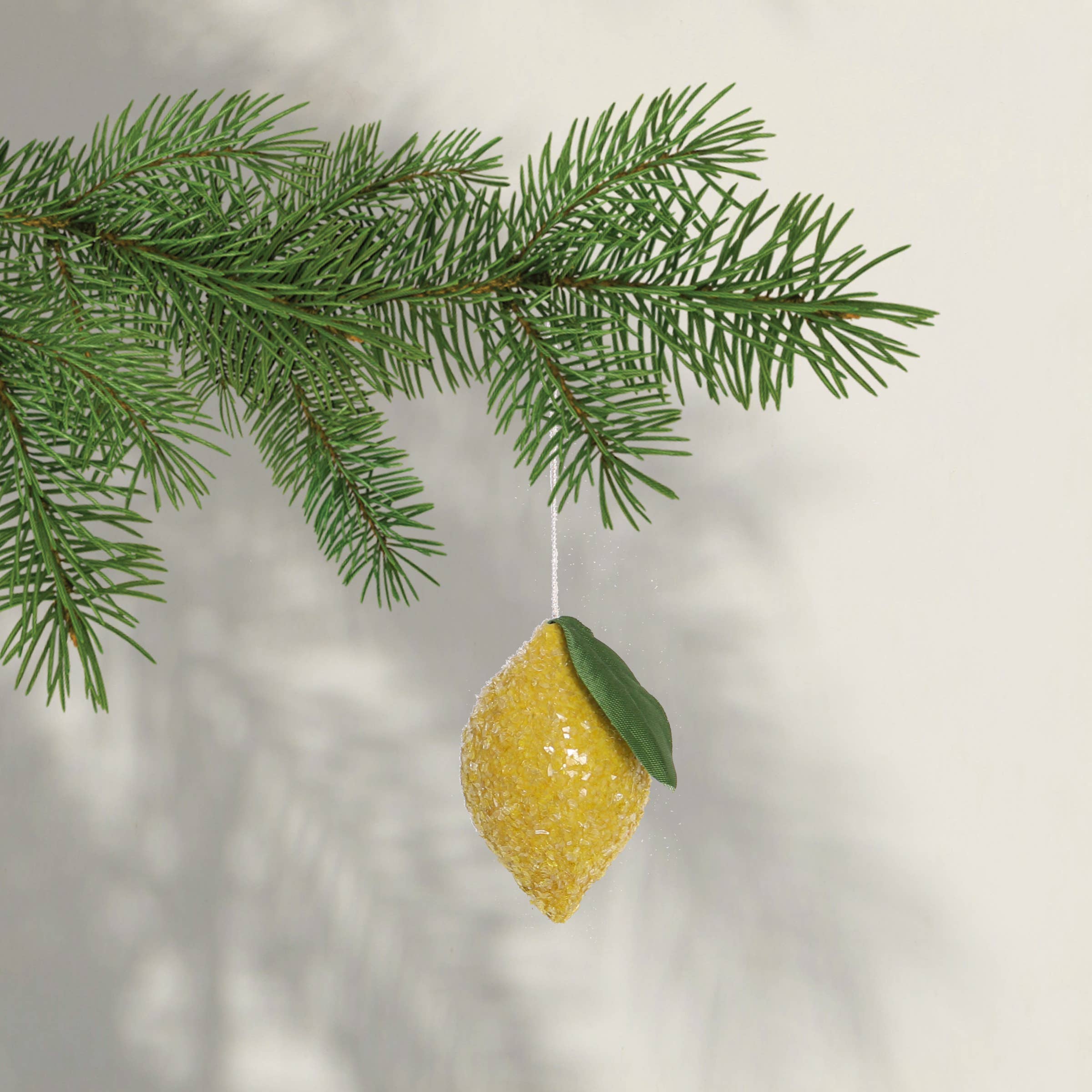 Decorative lemon ornament with a green leaf hanging from a pine branch against a blurred background