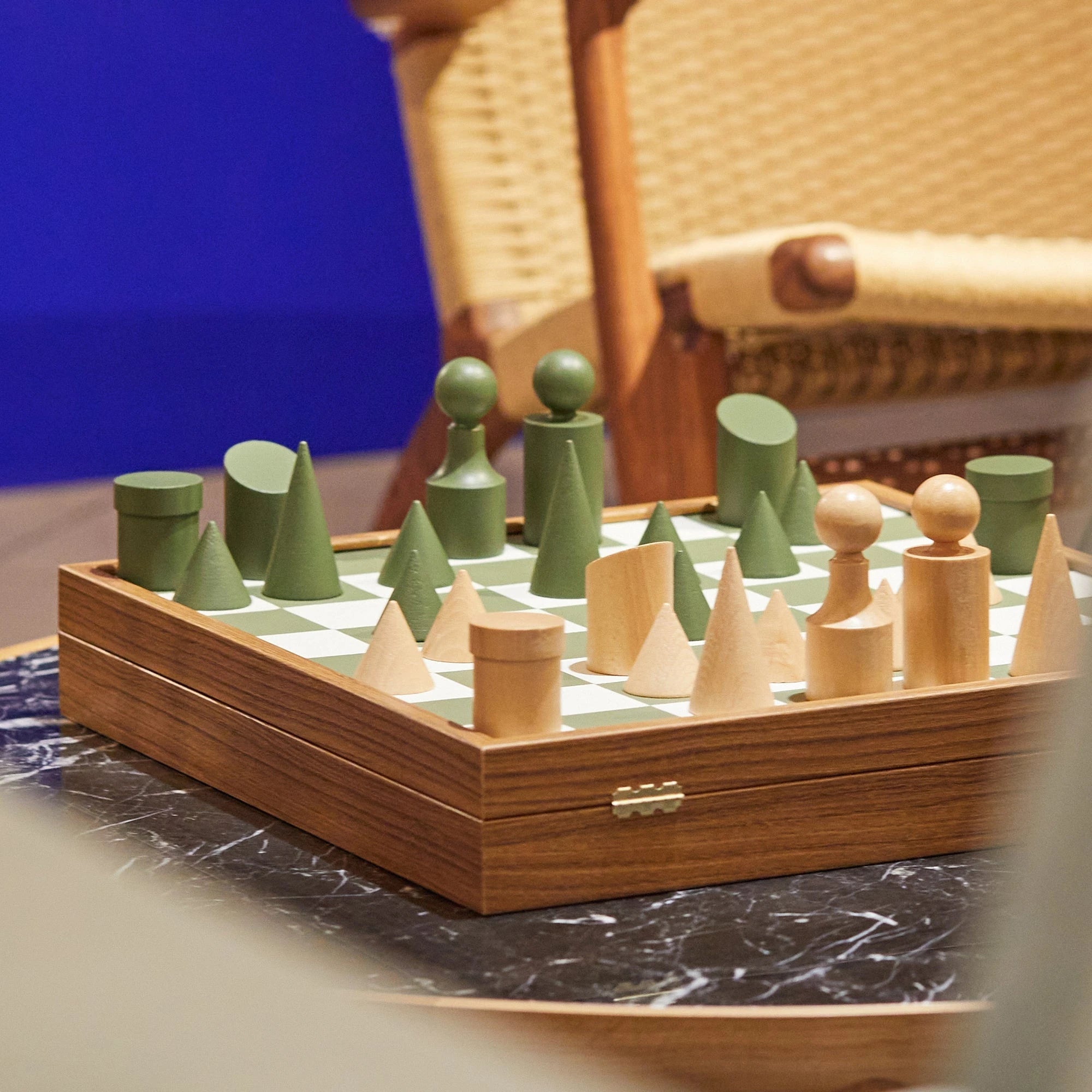 Milos Wooden Chess Set by The Conran Shop in olivine green mid play, set up on a marble table in front of a blue wall and rattan chair