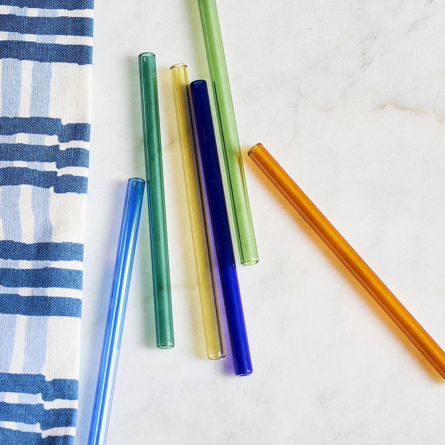 Santorini Set of 6 Glass Cocktail Straws by The Conran Shop on a white marble surface next to a blue checked towel