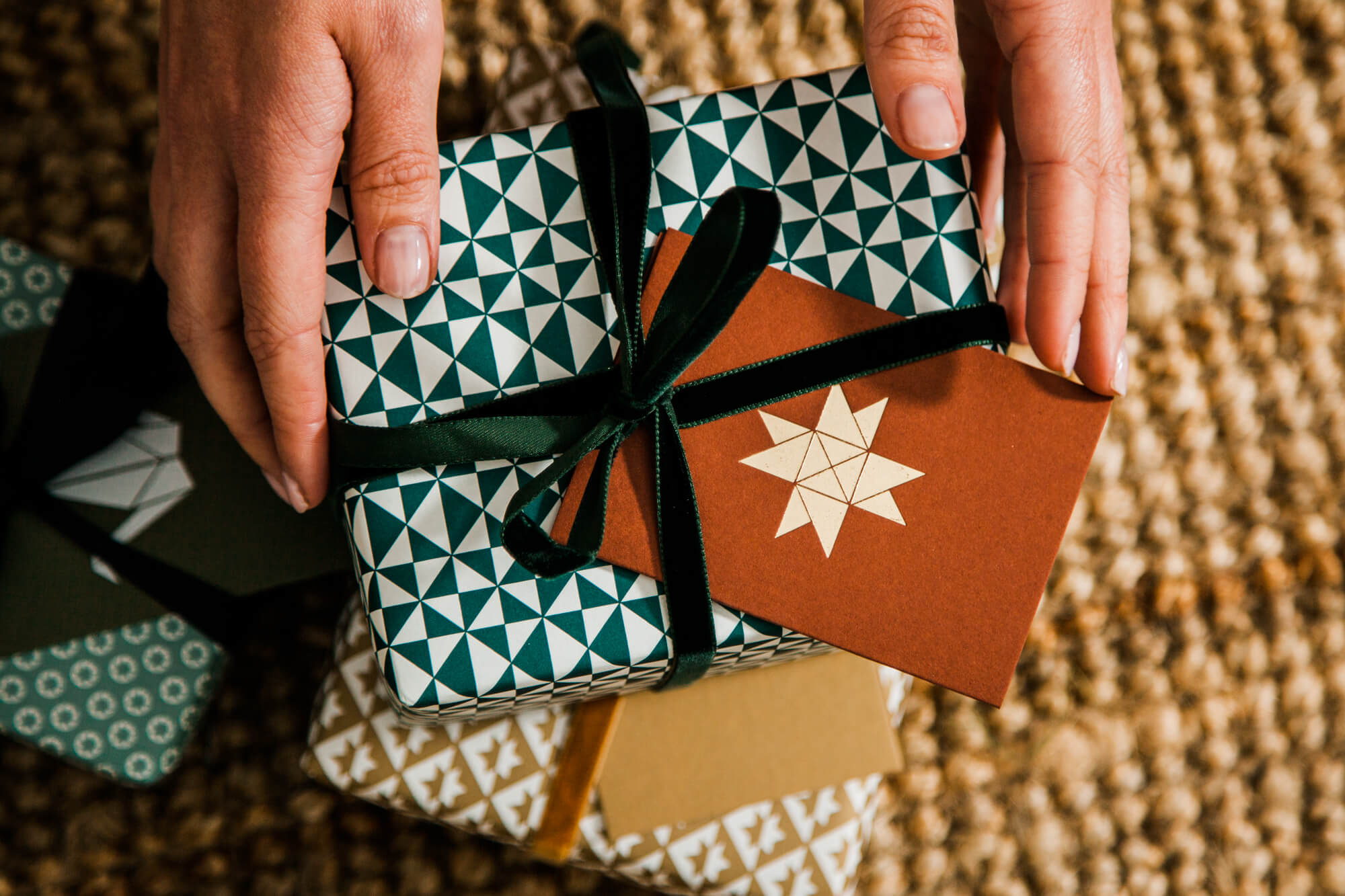 A pair of hands sliding a geometrically designed red Christmas card under the velvet ribbon on a wrapped present