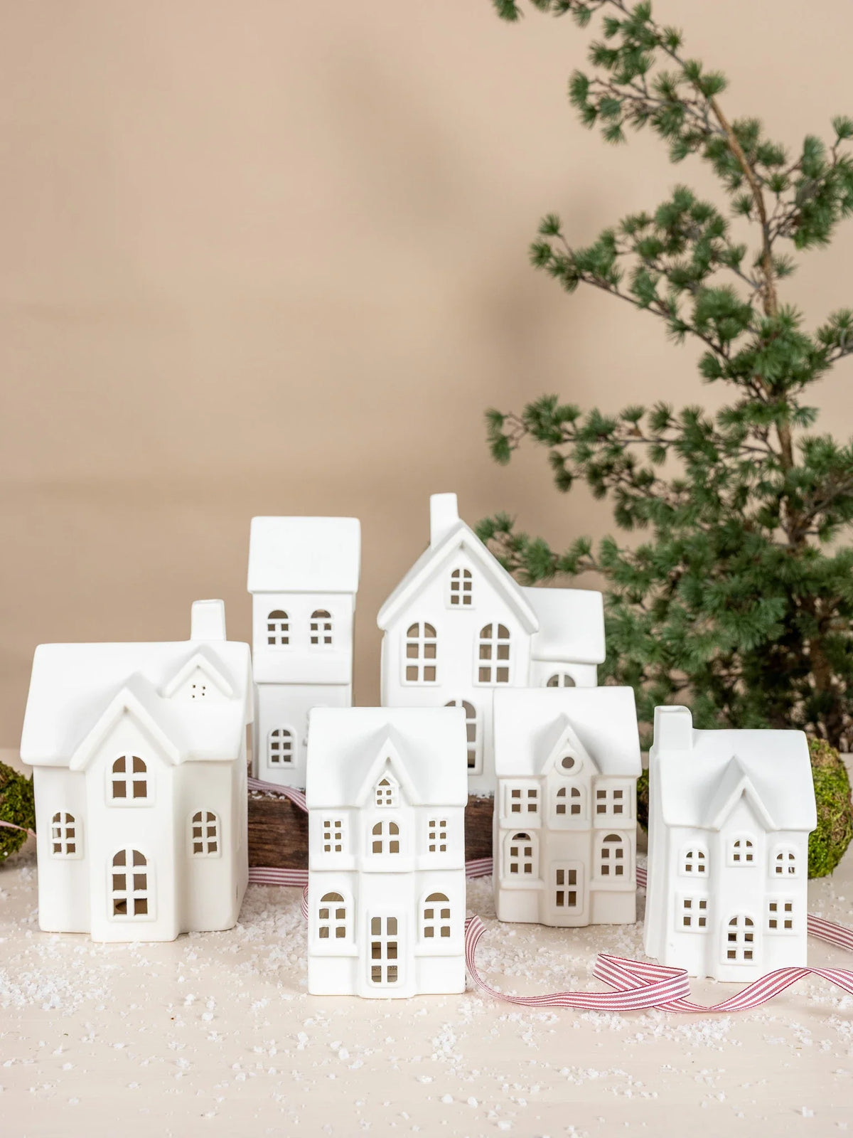 A set of small white ceramic Christmas Byn house ornaments by Storefactory with cutout windows, arranged on a light surface with a red-and-white striped ribbon and a blurred evergreen branch in the background.