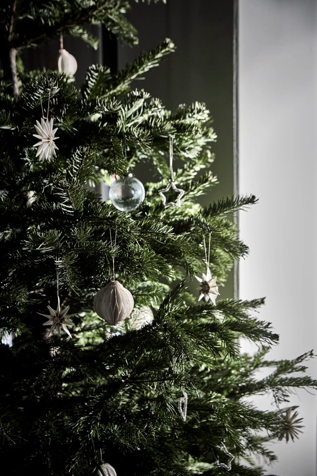 A close-up of a dark green Christmas tree decorated with matte silver and white House Doctor ornaments, including star and teardrop shapes, set against a dark and light grey background.