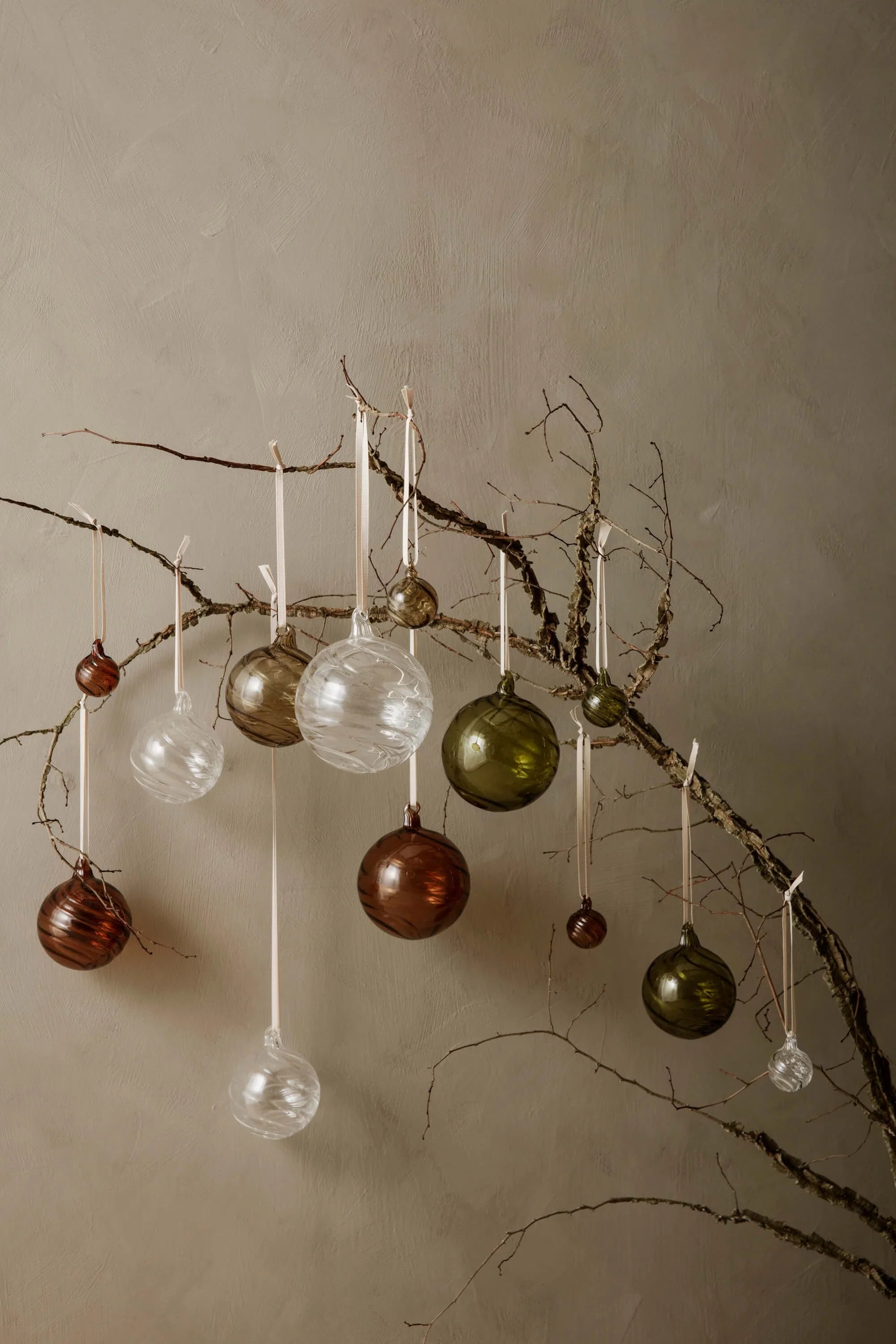 A series of glass festive baubles in various sizes and colours hanging from a single branch against a beige wall