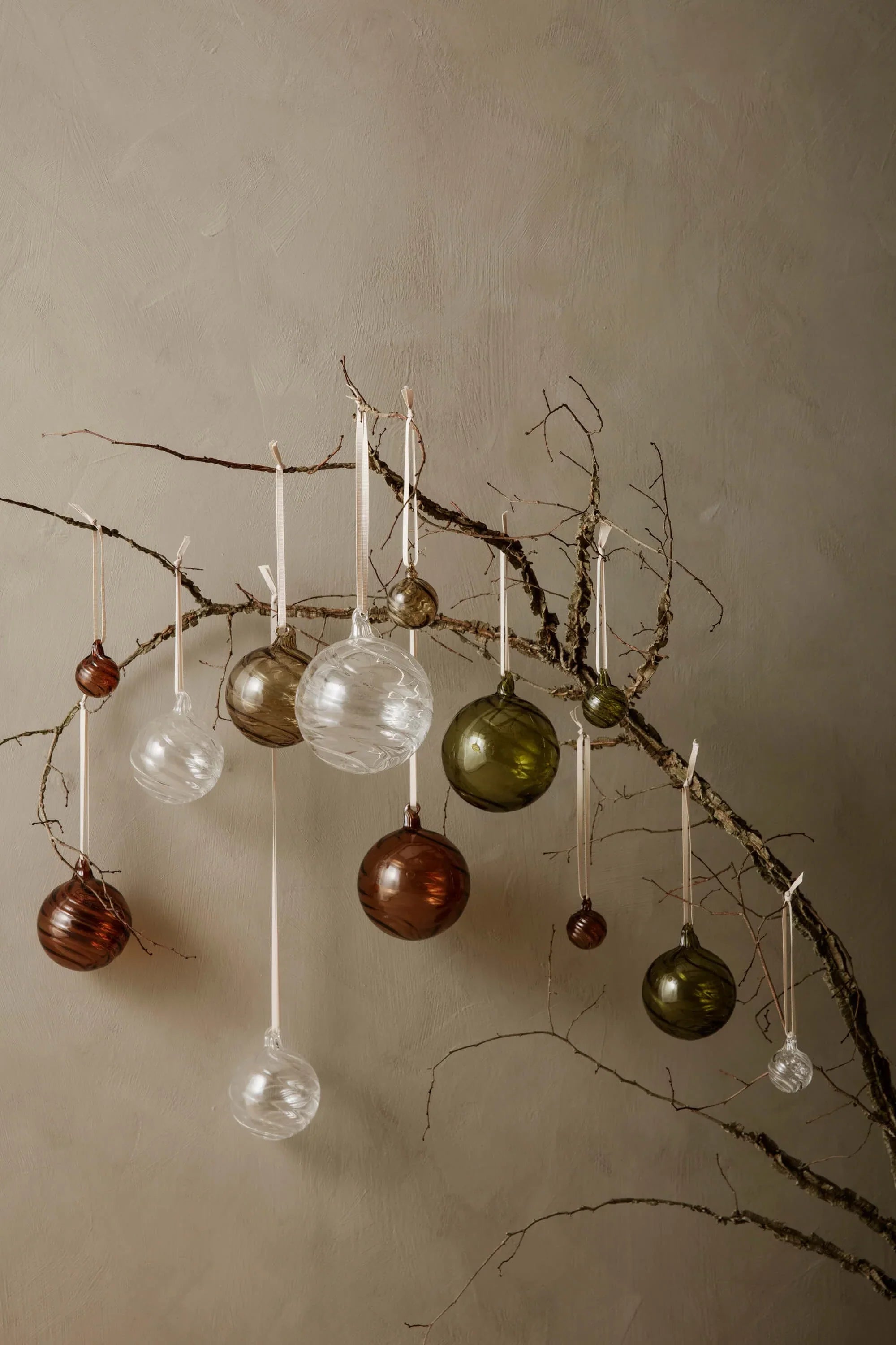 Christmas decorations : A series of glass festive baubles in various sizes and colours hanging from a single branch against a beige wall