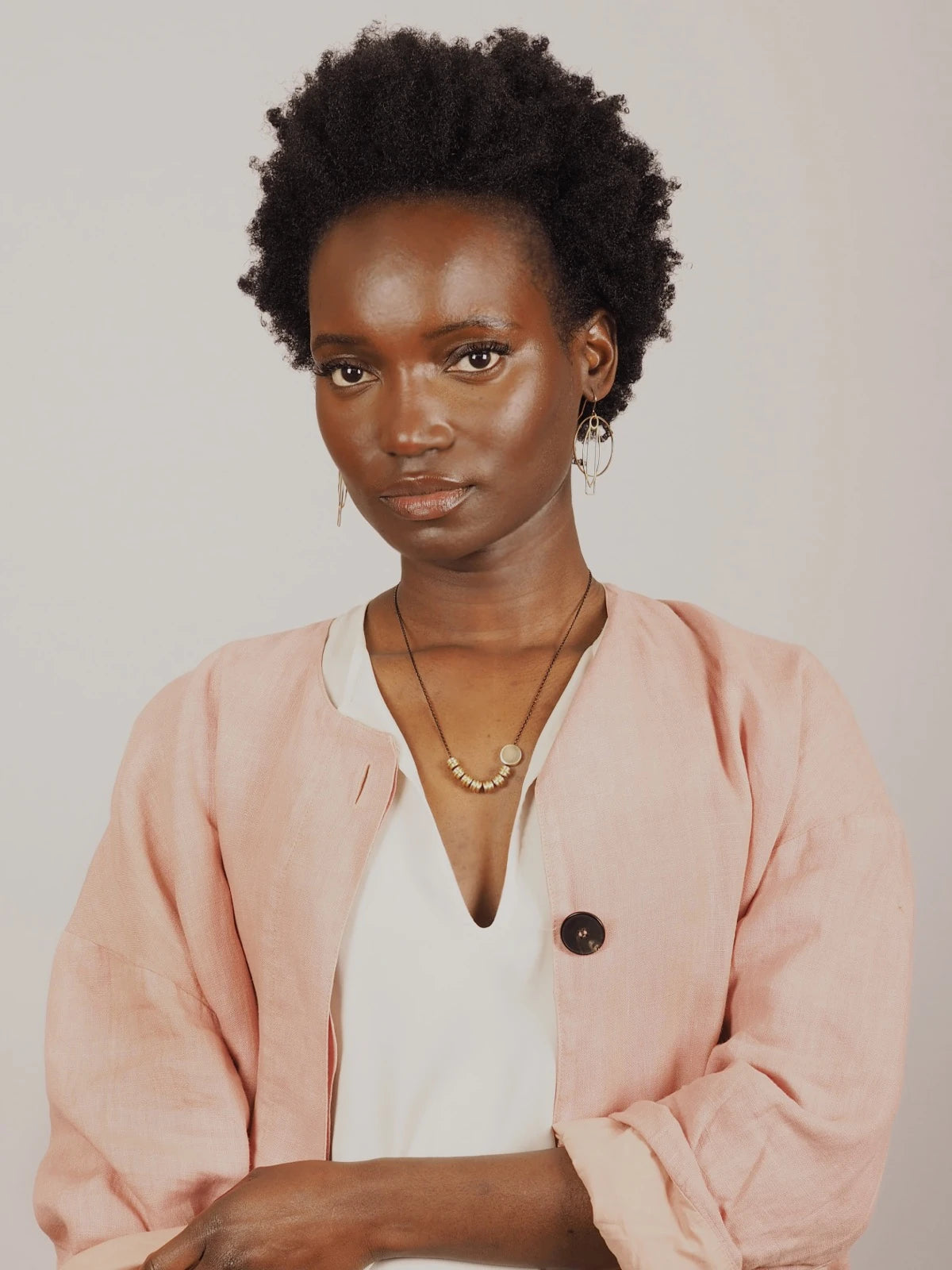 A woman with short curly black hair wearing a light pink linen jacket over a white top and Nucleo Solid Brass Totems and Brass Disc Necklace by brass+bold, photographed against a plain light grey background.